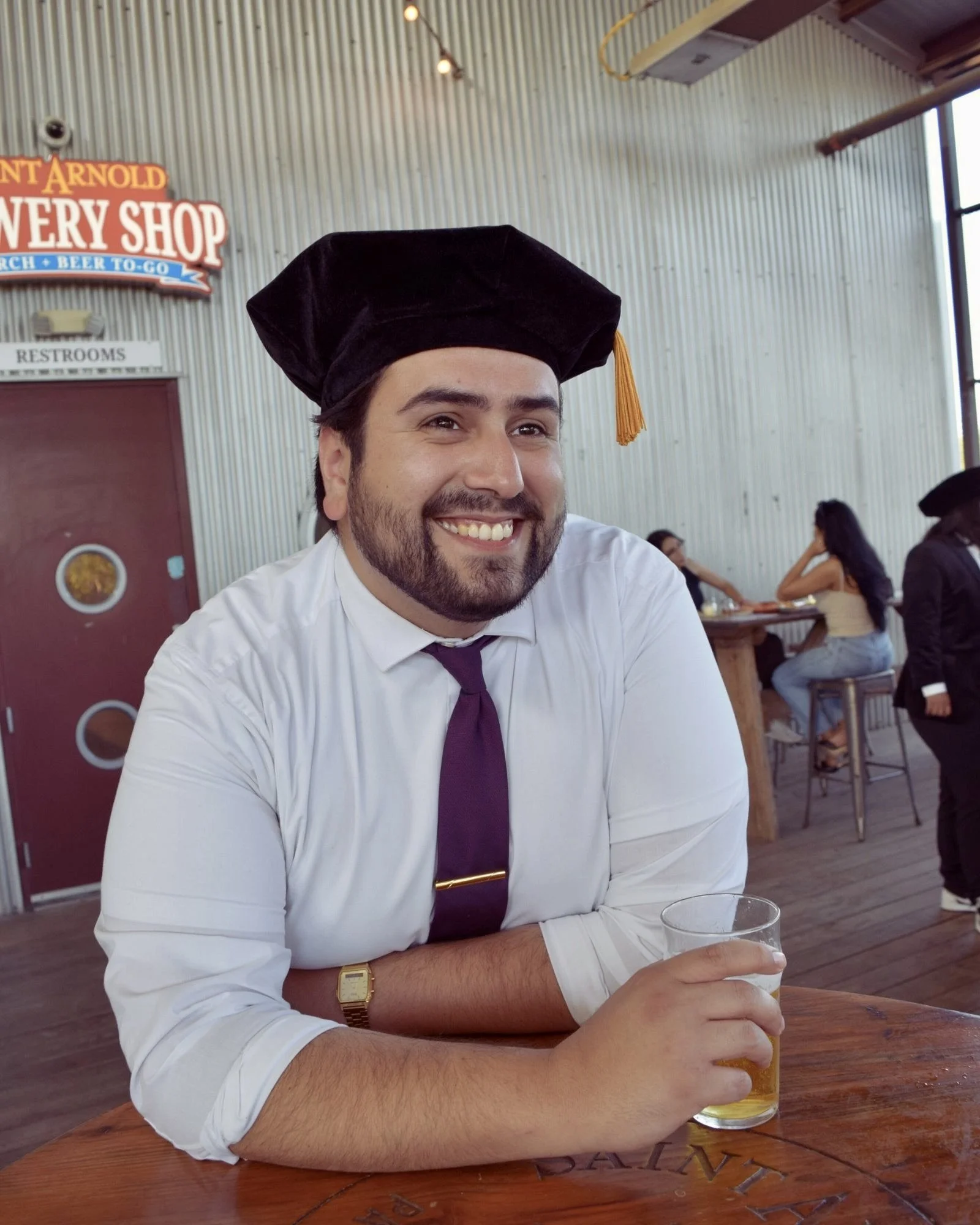 Man in a white shirt with rolled-up sleeves, wearing a purple tie and a black graduation cap, smiling while holding a glass of beer at a wooden table.