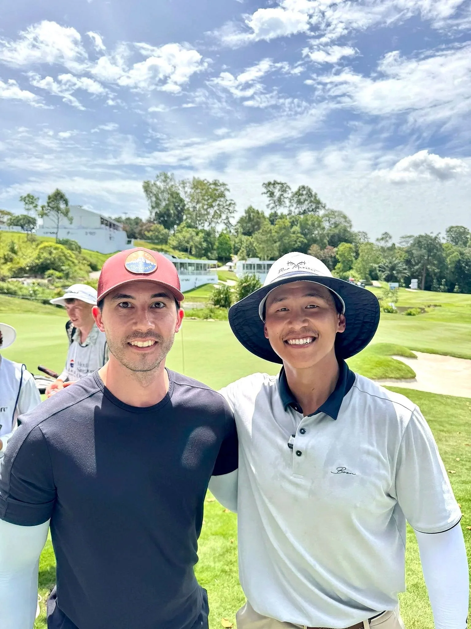 Two men standing together on a golf course, smiling, with a clear blue sky and trees in the background.