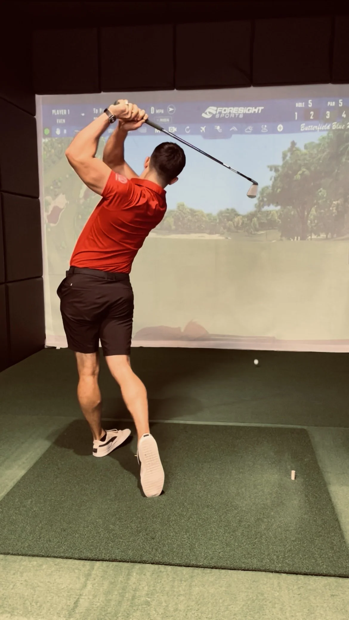 A man in a red shirt and black shorts practicing golf swings in a golf simulator room, with an indoor green screen displaying a golf course.