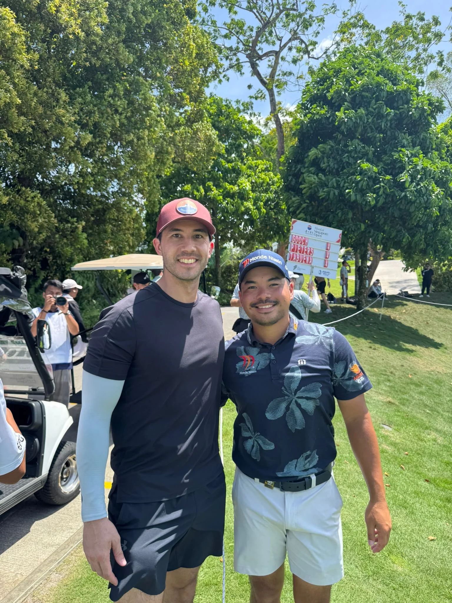 Two men smiling and posing for a photo on a golf course. One is wearing a black shirt, black shorts, and a red cap; the other is wearing a navy blue Hawaii-themed golf shirt, white shorts, and a navy cap. There are trees, a golf cart, and a scoreboard in the background.