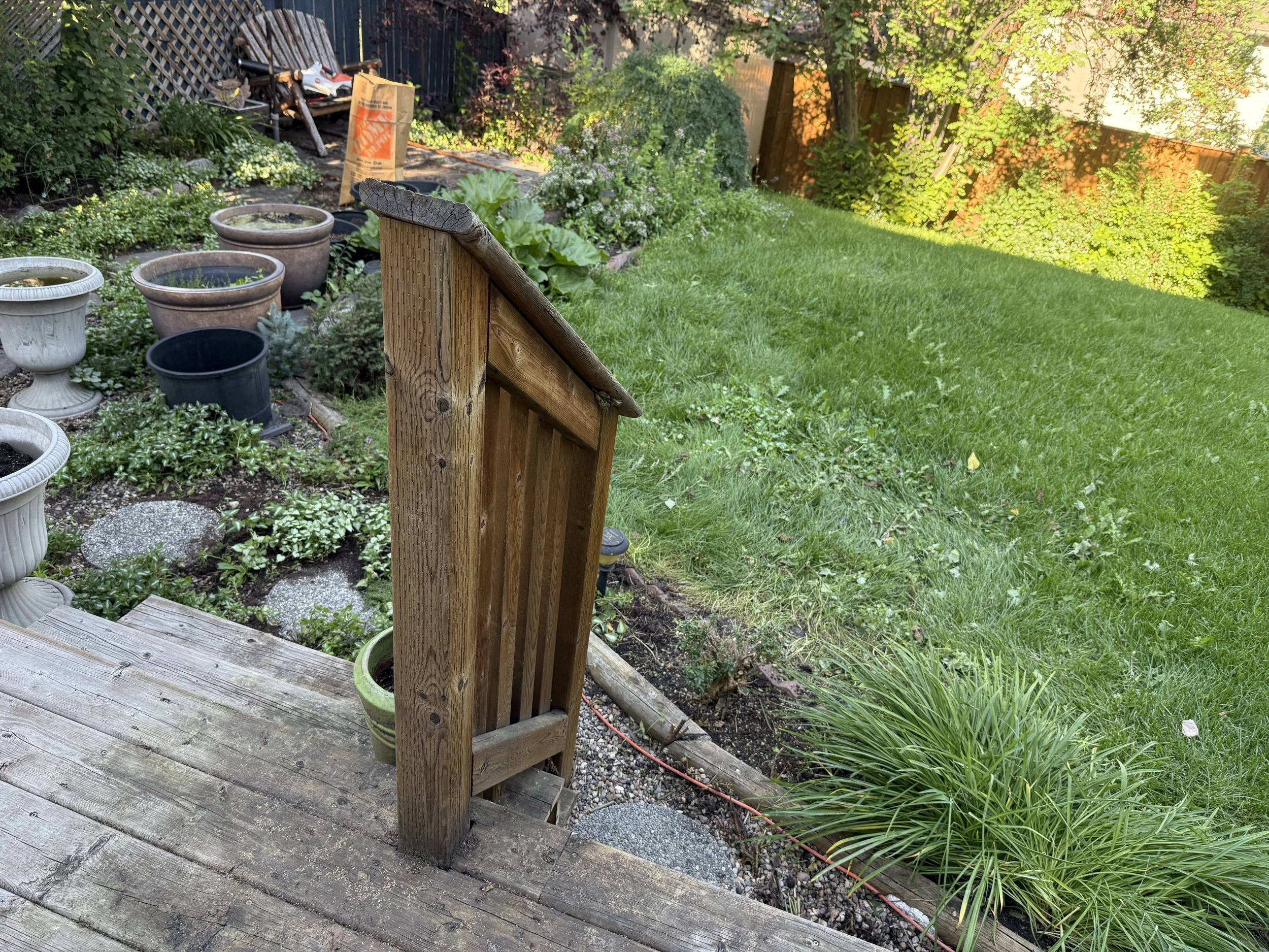 A backyard garden with potted plants, green grass, and a wooden deck with a railing. There are small flowering plants and trees along the fence line from garden maintenance in Calgary