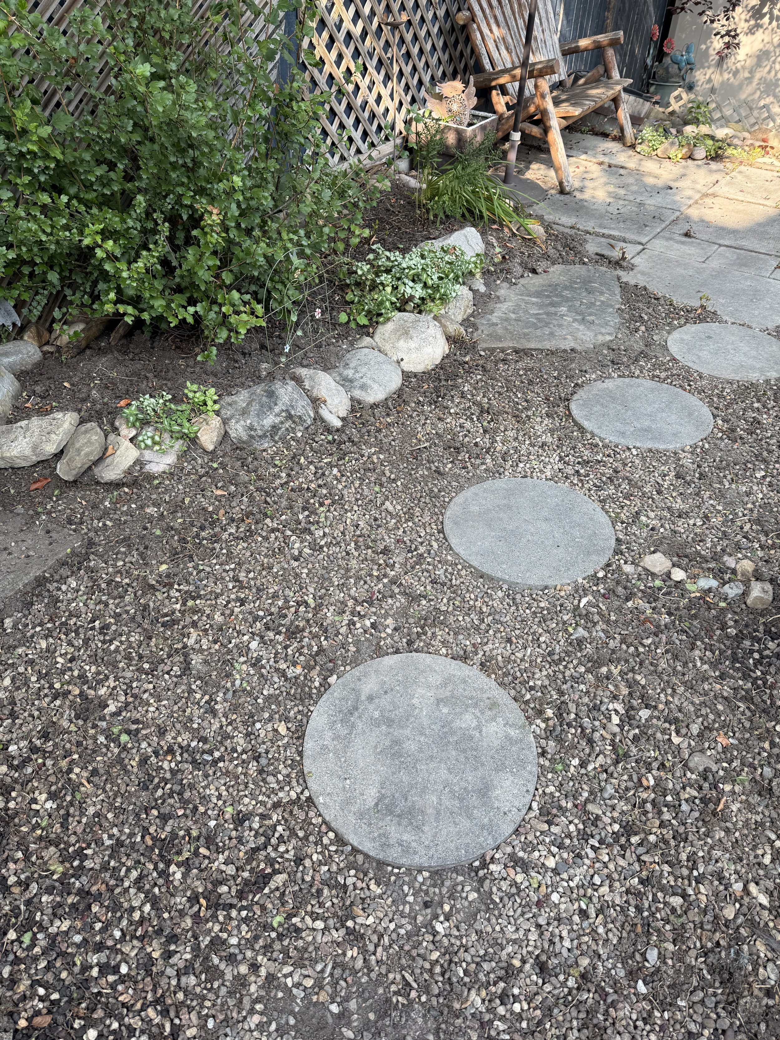 A backyard garden with a pebble pathway, rocks along the edge of a flower bed, and a rustic wooden bench. There are various plants and decorative garden ornaments, including a butterfly and owl figurine. Completed during a weekly garden maintenance