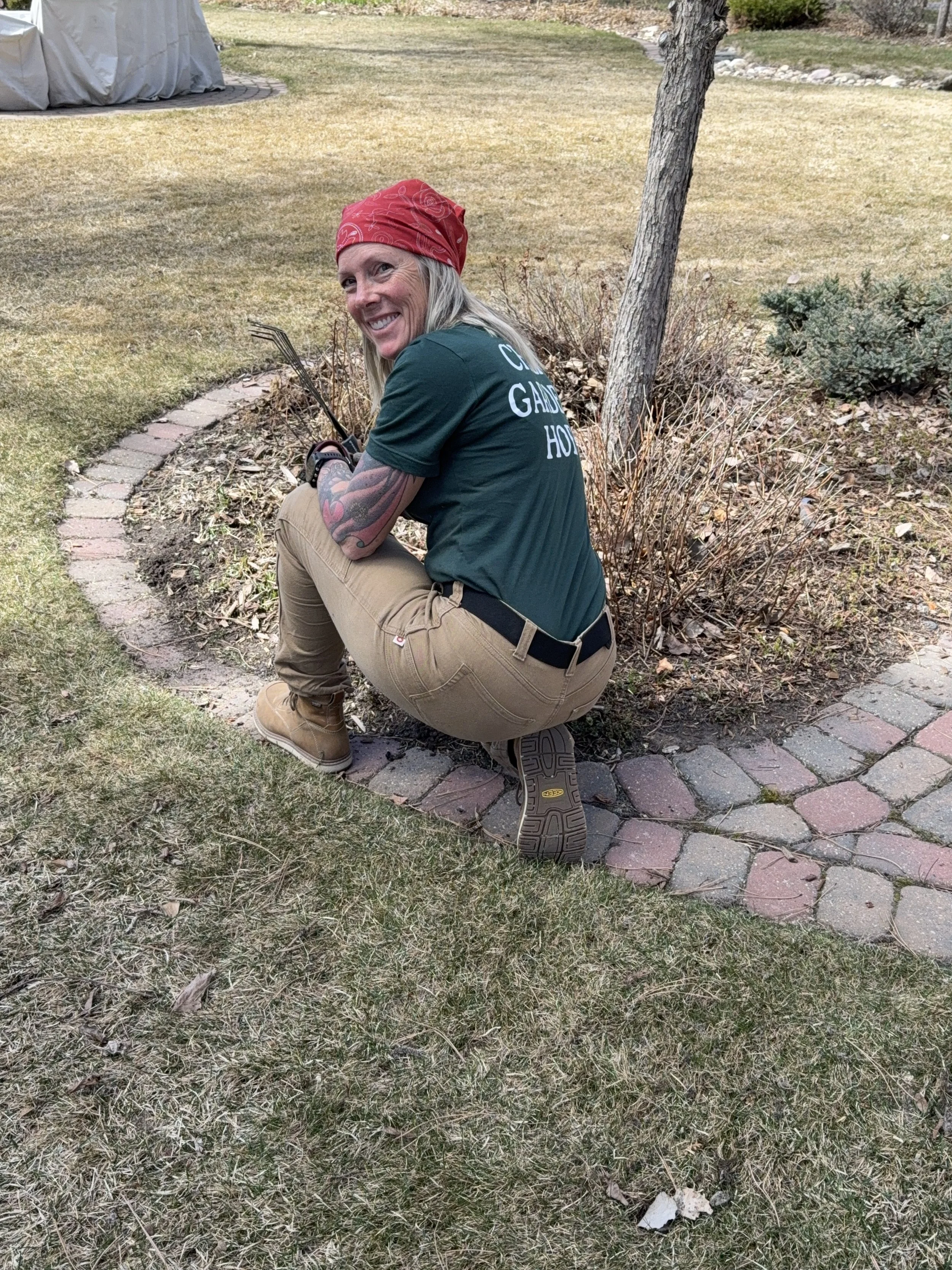A woman, Jen, who runs a garden maintenance business in Calgary with tattoos on her arm, wearing a red bandana, green shirt, tan pants, and tan boots, is squatting on a brick pathway in a yard with grass, trees, and bushes, smiling at the camera.