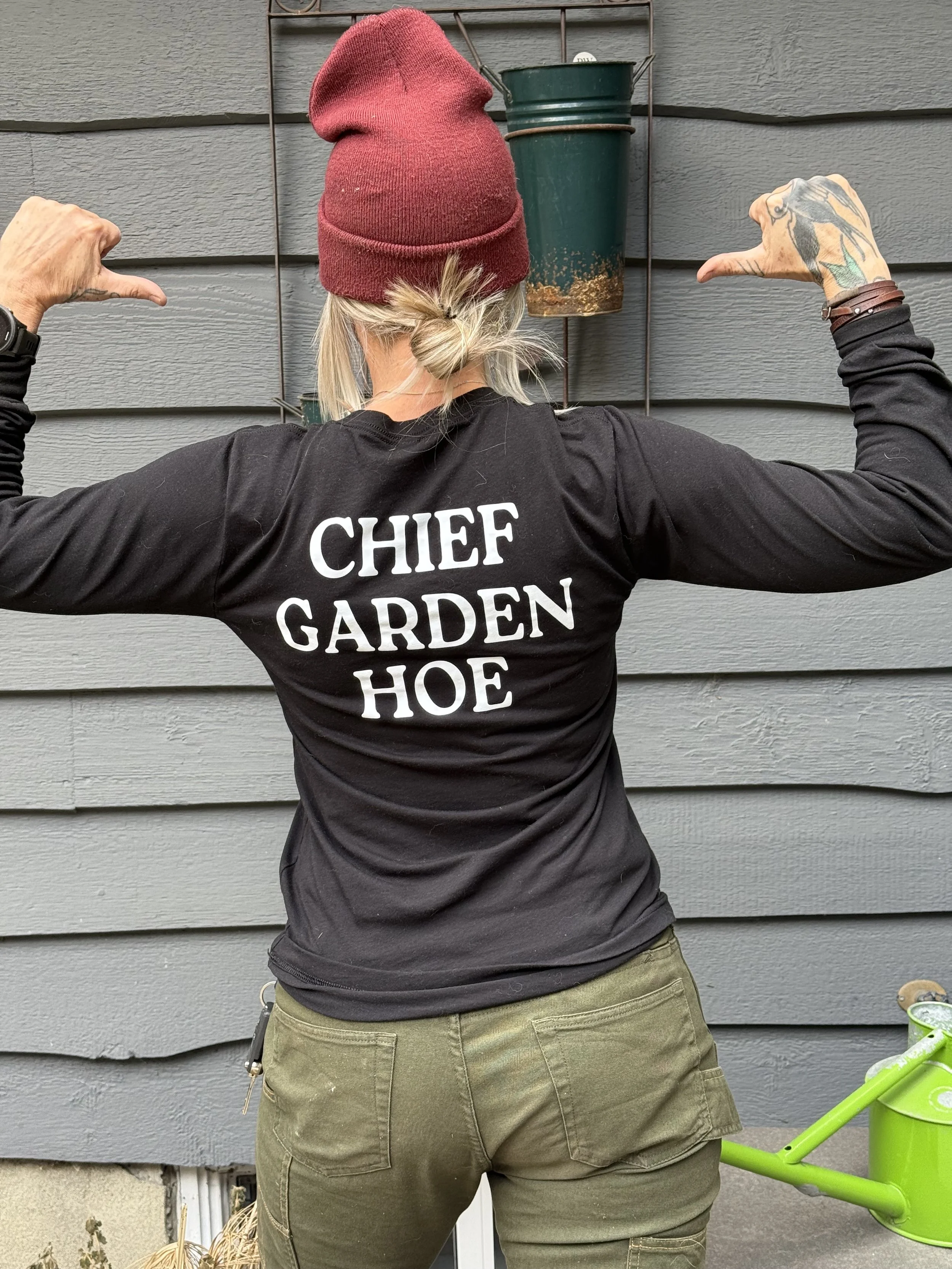 Back of Jen, a woman, who owns a garden maintenance business in Calgary, wearing a red beanie, black long sleeve shirt with white text that reads 'CHIEF GARDEN HOE,' and green cargo pants, pointing at a green hanging flowerpot on a gray house wall.