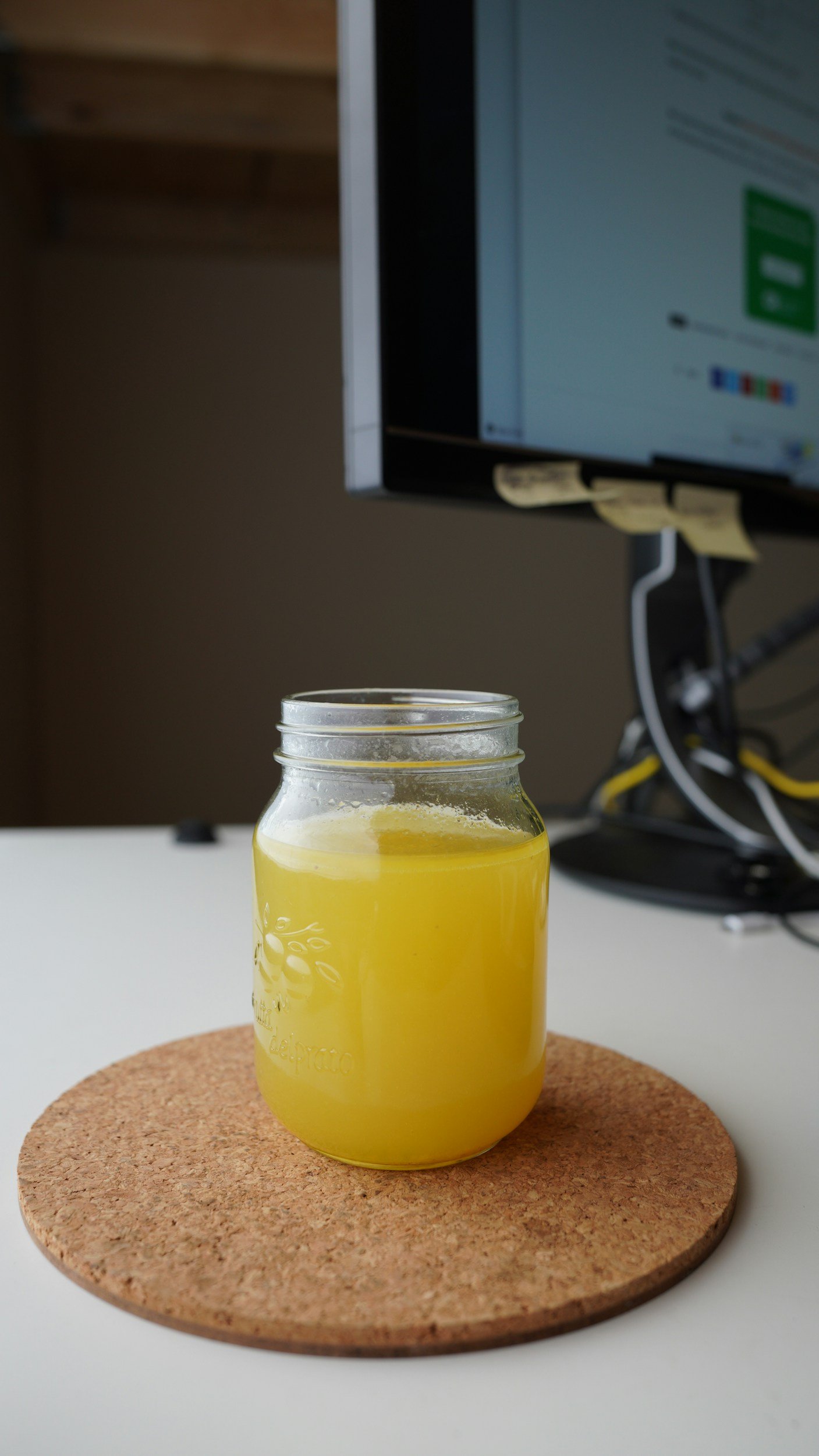 A jar filled with yellow liquid placed on a cork coaster on a white table, with a computer monitor and cables in the background.