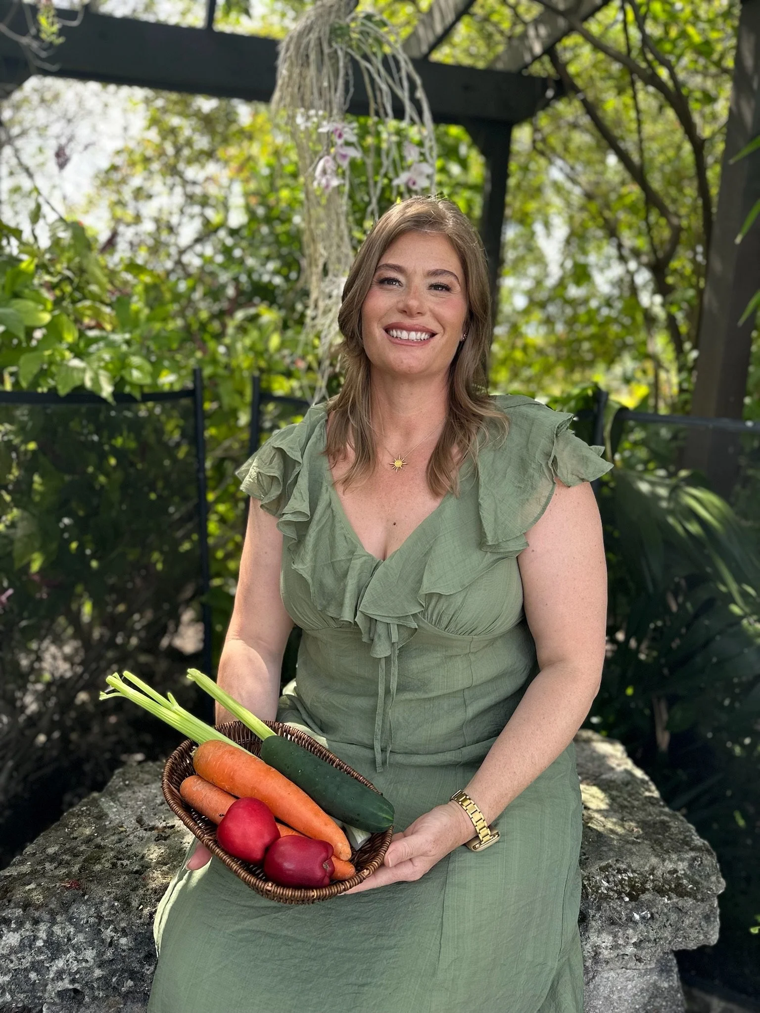 A woman with light brown hair, wearing a green dress and a gold watch, is holding a basket of carrots and red peppers outdoors in a garden.