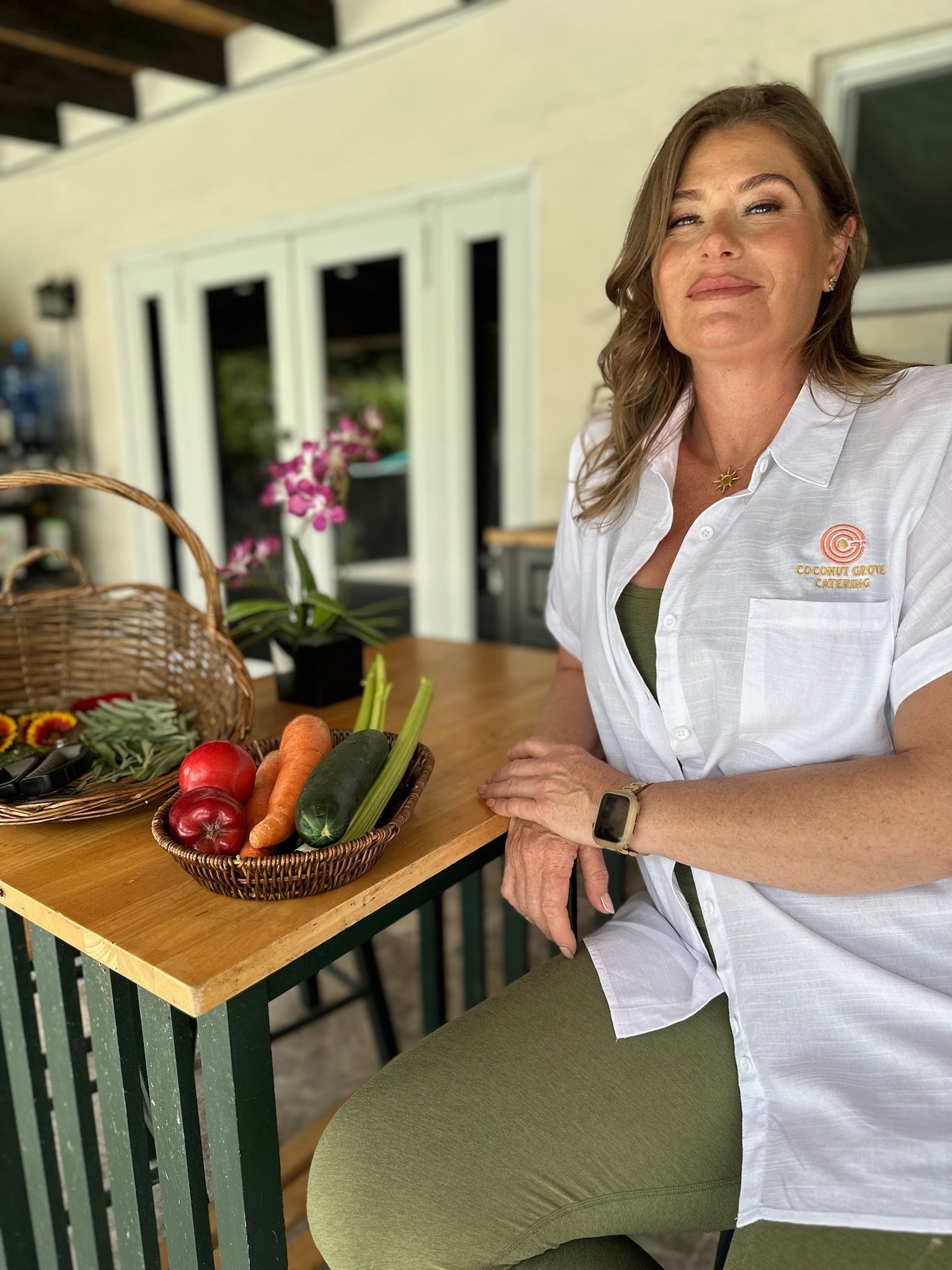 A woman sitting at a wooden table with a basket of fresh vegetables, including carrots, zucchini, red onions, and celery, in front of her. She is wearing a white shirt with a logo that reads 'Coconut Grove Catering' and a gold necklace, and she is smiling slightly while posing for the photo.