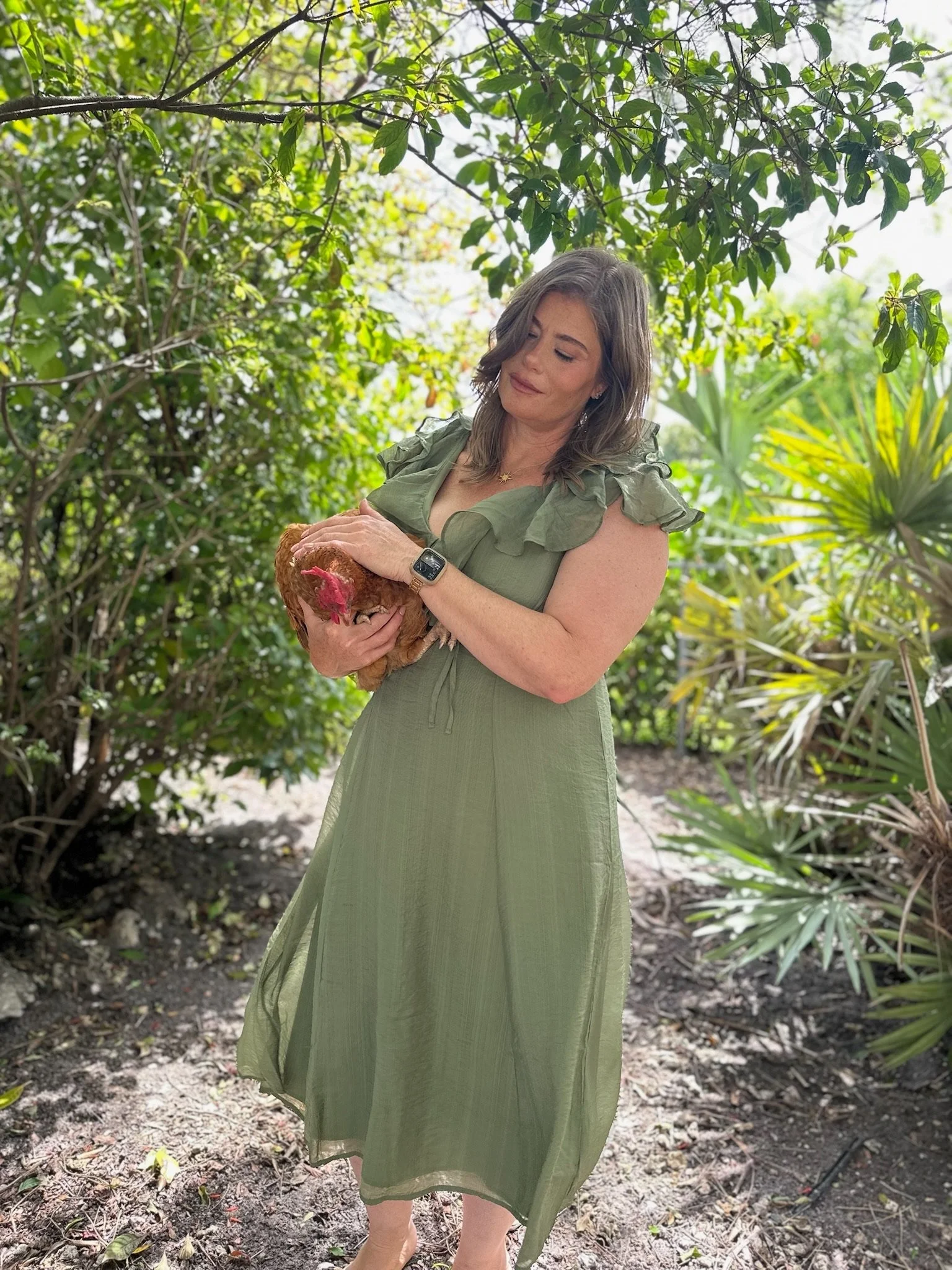 A woman wearing a green dress stands outdoors among green foliage, holding a brown chicken.