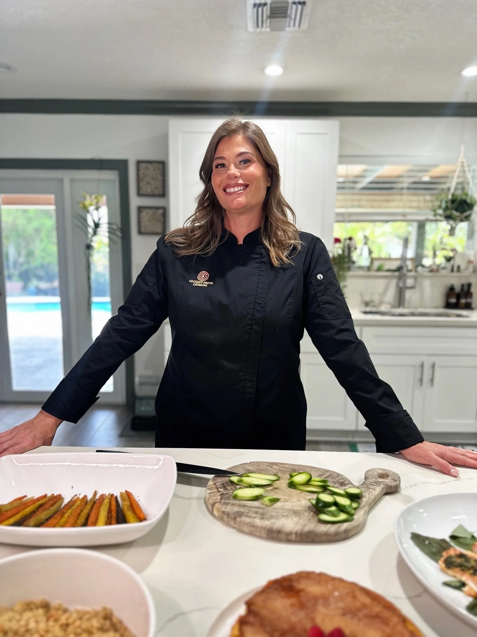 A woman in a black chef's coat standing in a modern kitchen with various dishes on the counter, including sliced cucumbers, roasted vegetables, and other plated food items.