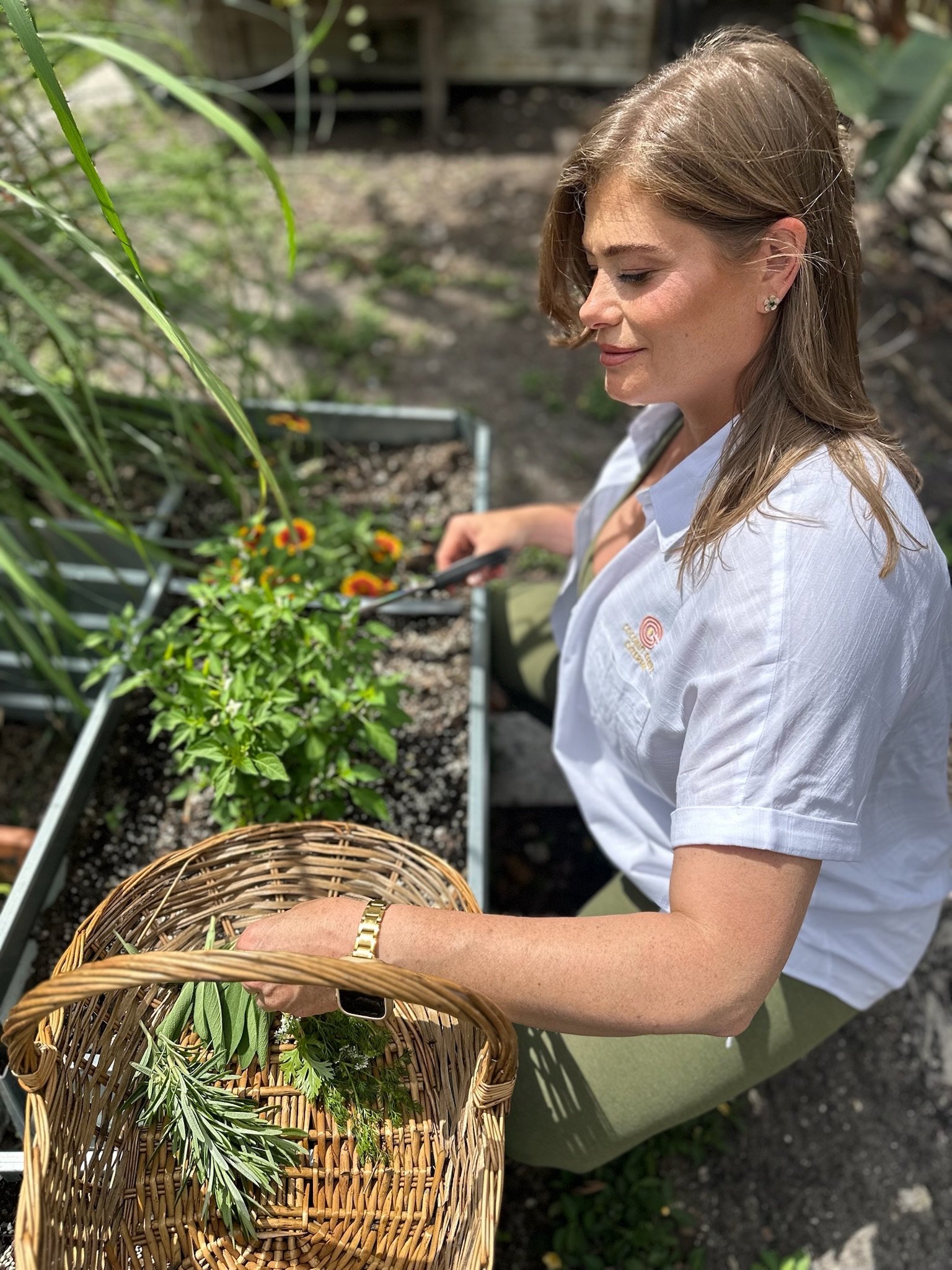 A woman with light brown hair, earrings, and a white shirt, sitting outdoors, holds a wicker basket filled with green plants and herbs, with gardening tools and plants around her.