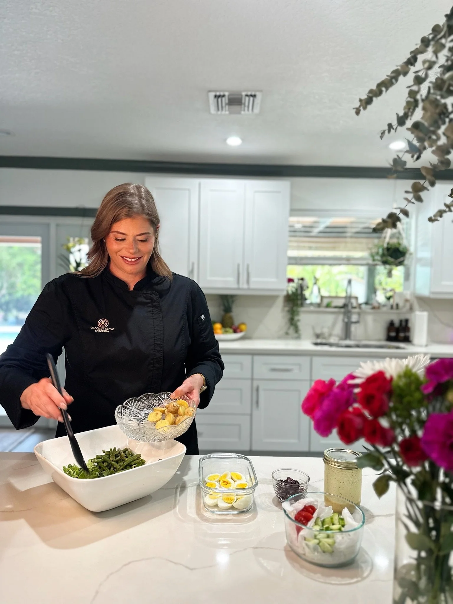 A woman in a black chef's jacket preparing a salad in a bright, modern kitchen with white cabinets and large windows. Various bowls with vegetables and toppings are on the counter, and there are flowers in the foreground.