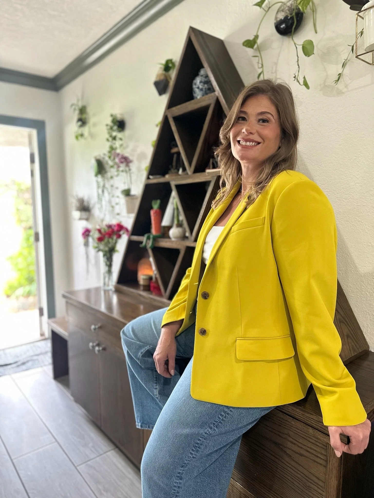 A woman wearing a bright yellow blazer and blue jeans, sitting on a wooden bench indoors, with a light-colored wall behind her decorated with hanging potted plants and a triangular wooden shelf with decorative items.