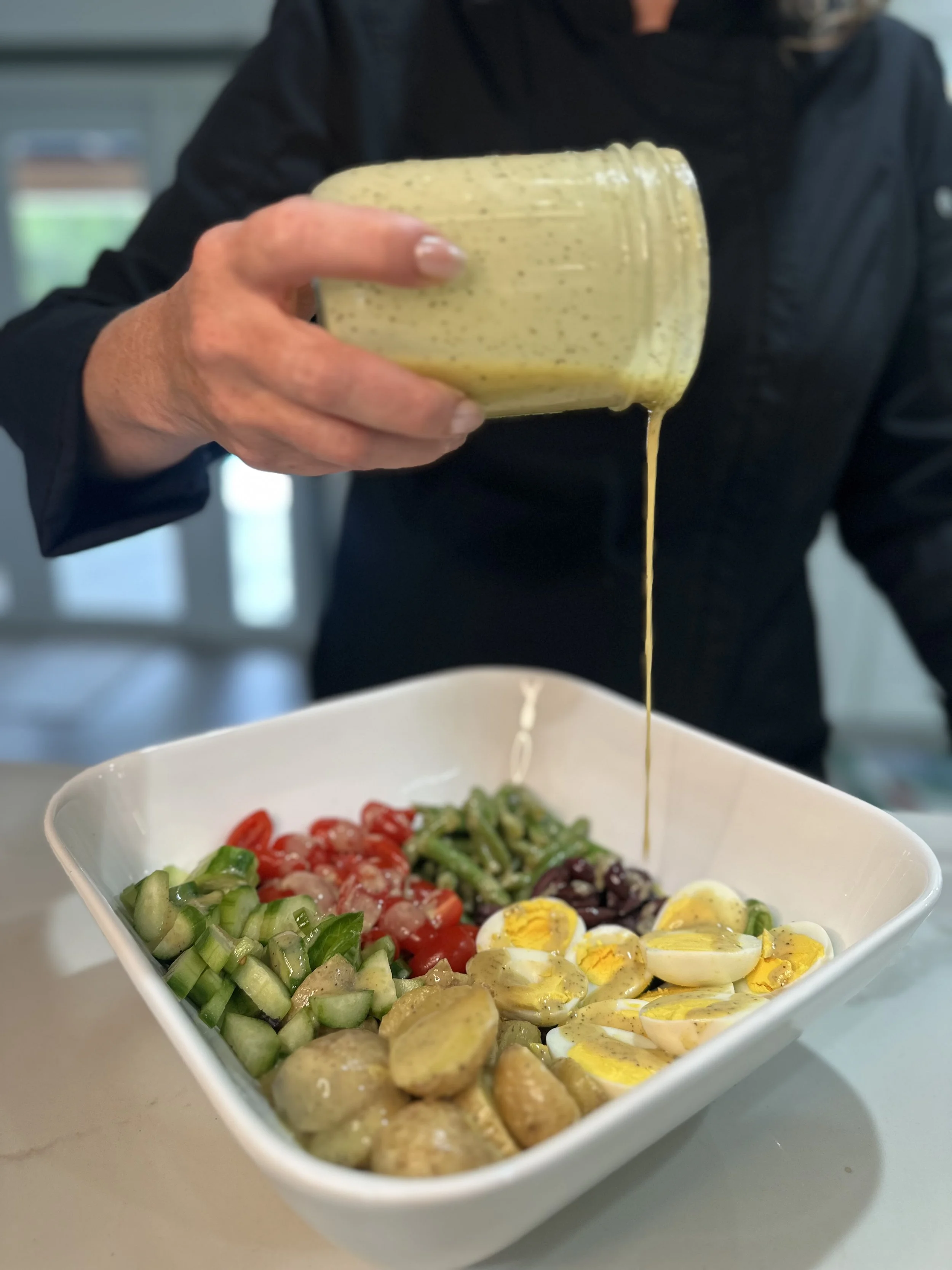 Person pouring salad dressing over a bowl of salad containing hard-boiled eggs, cherry tomatoes, cucumbers, olives, and grilled chicken.