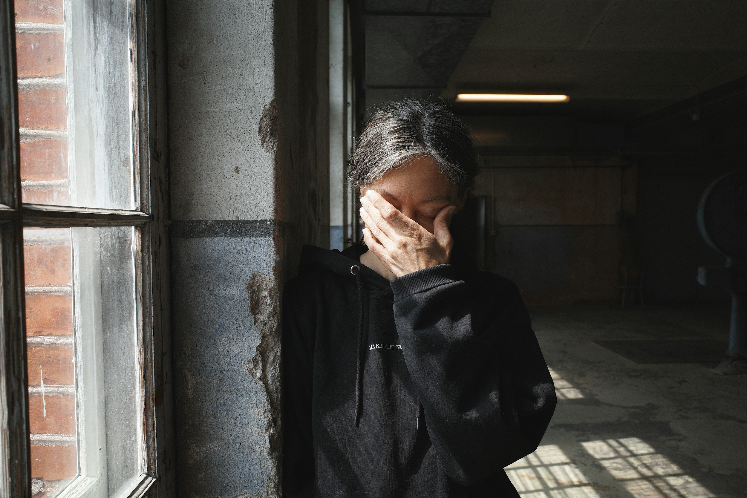 Une femme d'âge moyen se tient près d'une fenêtre, couvrant son visage avec sa main, dans un bâtiment en état de délabrement.