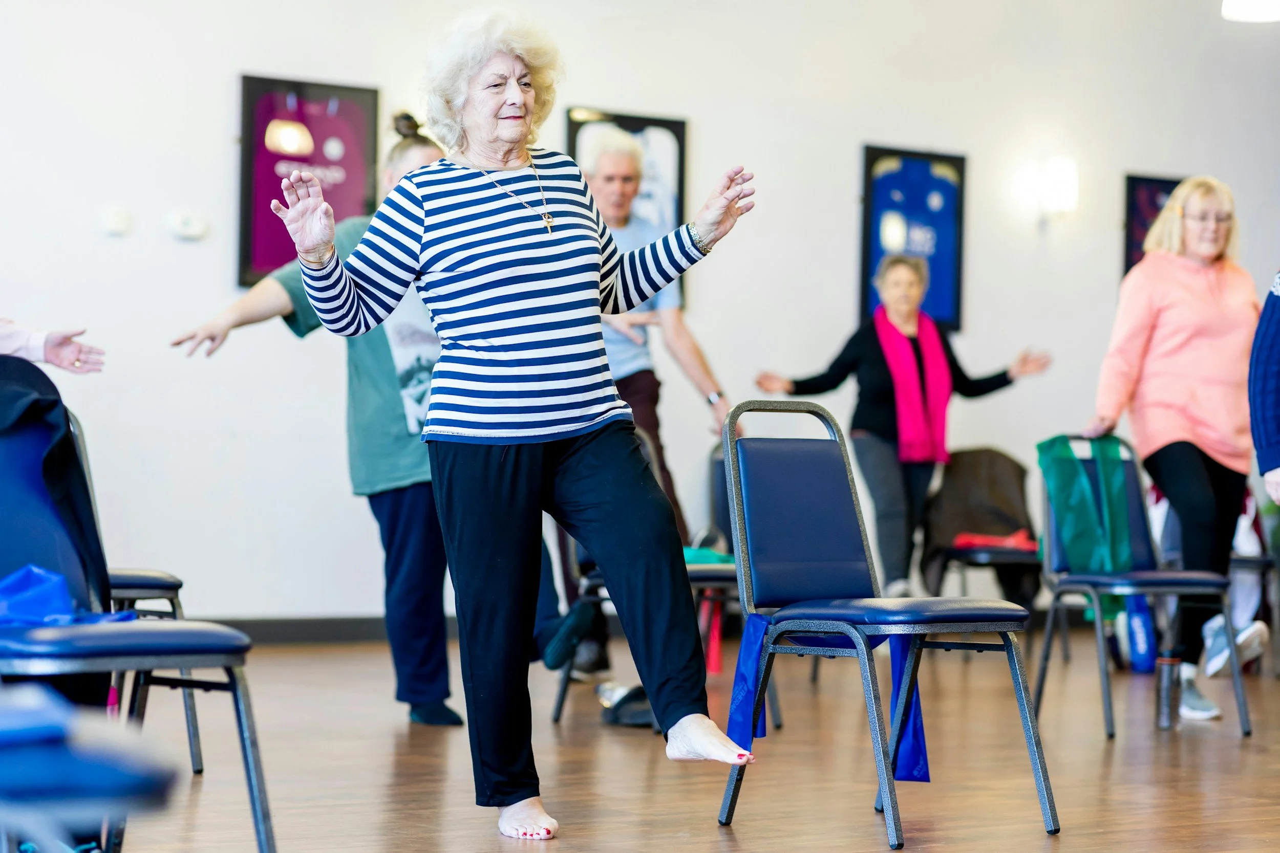 Une dame âgée participe à une séance de mouvement dans une salle de yoga ou de danse, entourée d'autres personnes également engagées dans des activités de mouvement, avec des chaises à proximité.