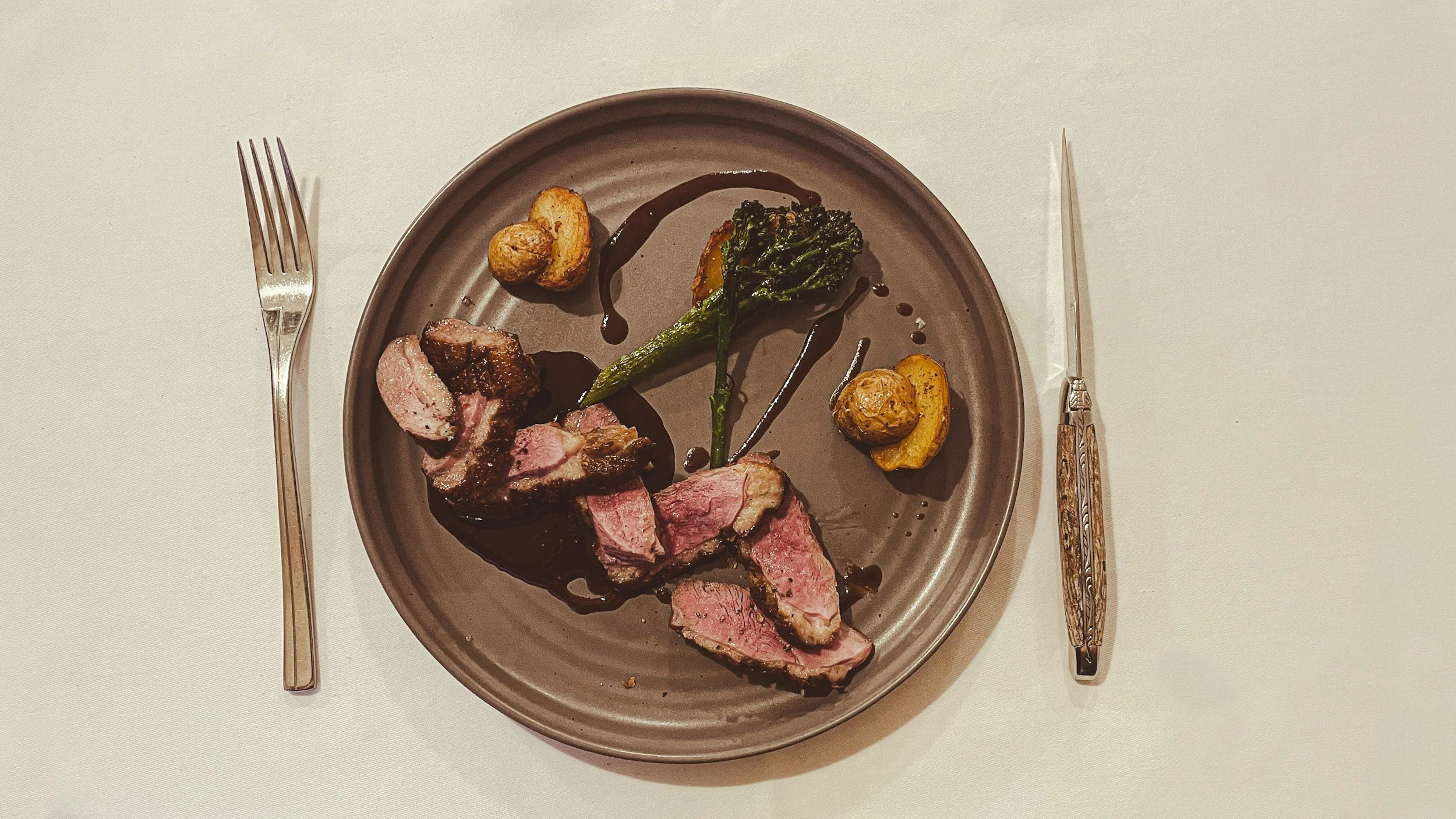 Assiette de filet de boeuf saignant avec légumes et sauce au vin, accompagnée de pommes de terre rôties, sur une table avec couverts.