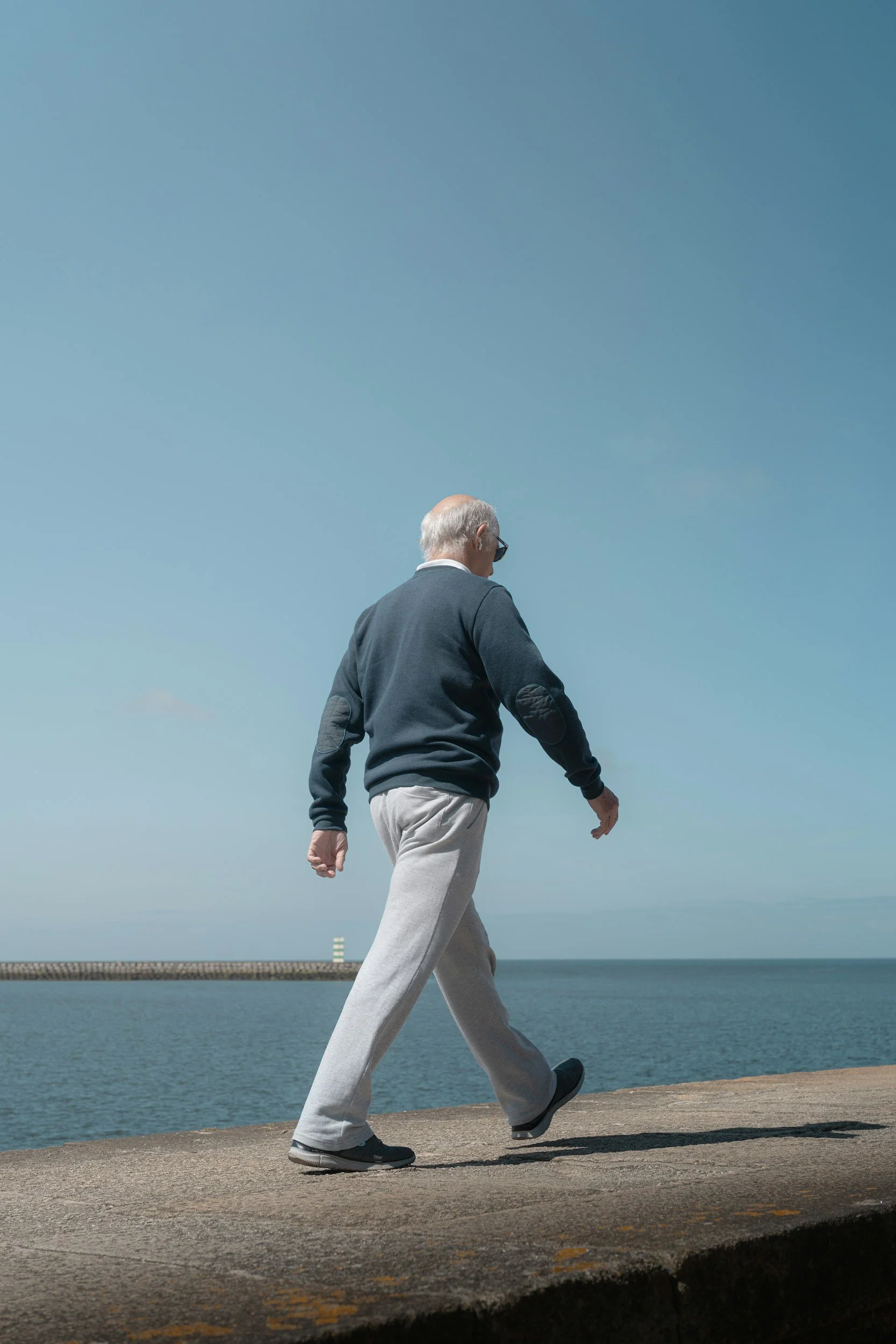 Un homme âgé marche le long d'une digue près de la mer, portant des vêtements décontractés et des lunettes de soleil.