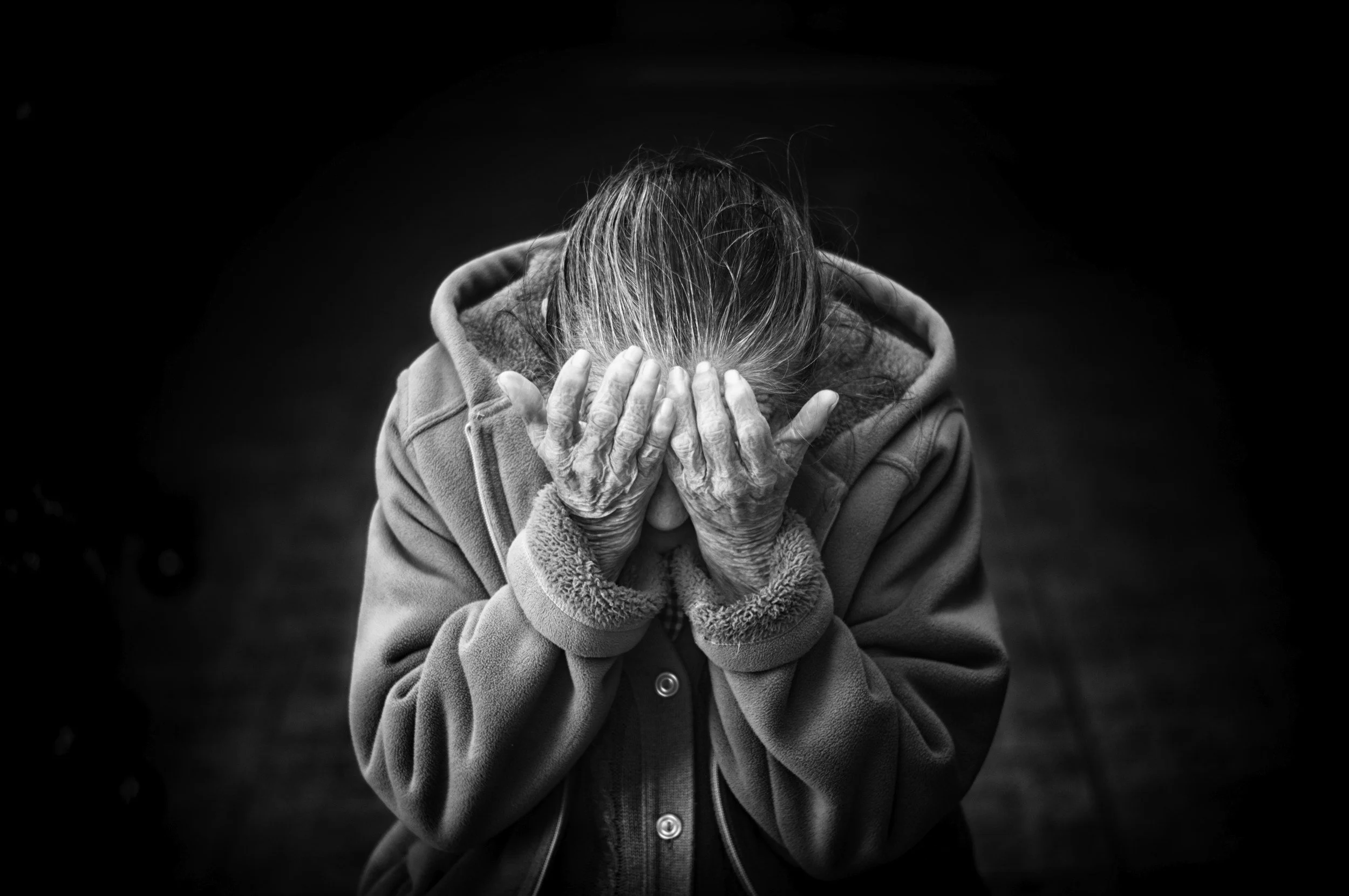 Une personne âgée en sweat à capuche, tête baissée, mains couvrant son visage, exprimant une détresse ou une tristesse. Photographie en noir et blanc avec fond sombre.
