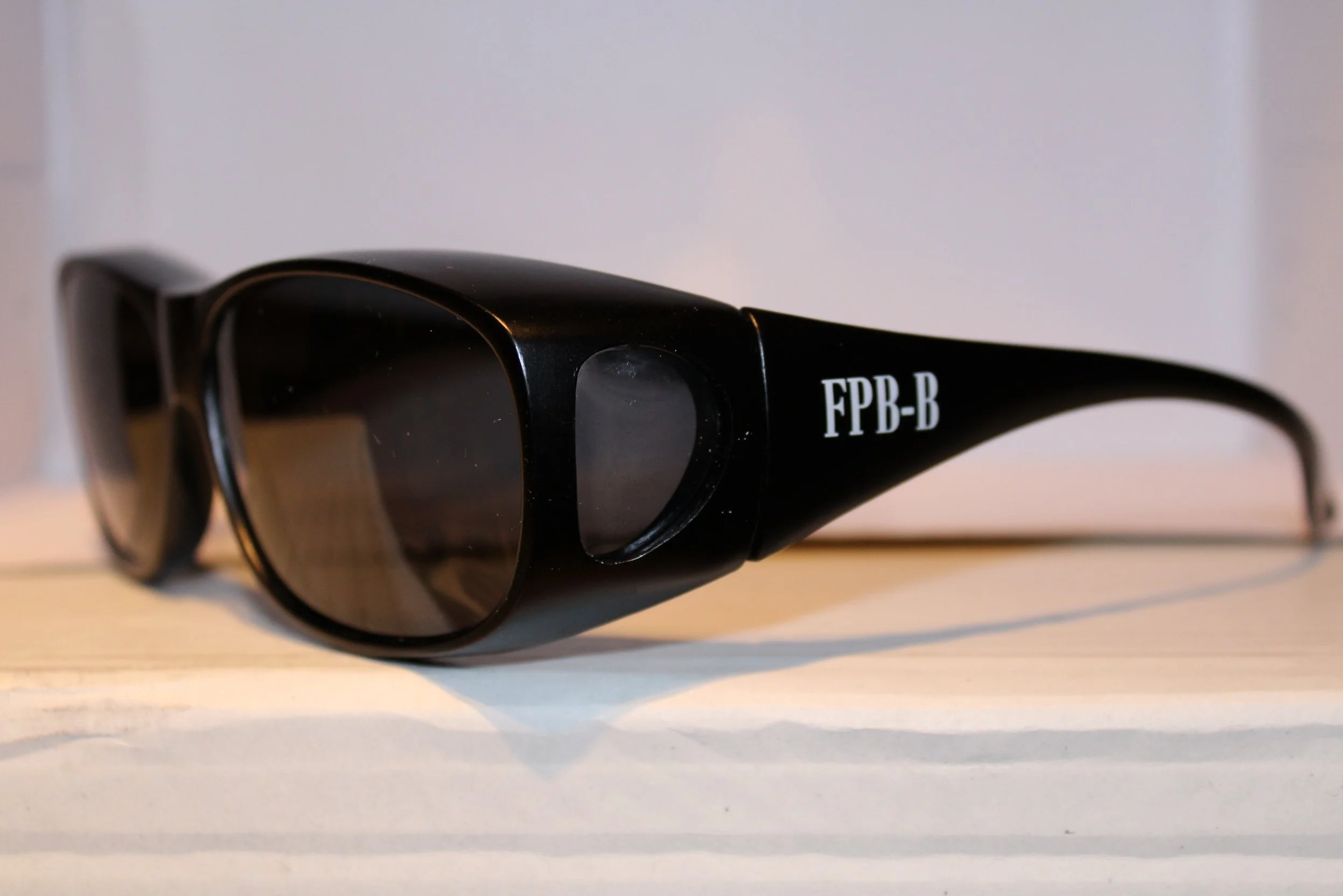 Lunettes de soleil noires avec le marquage 'FPB-B' sur la branche, posées sur une surface claire.