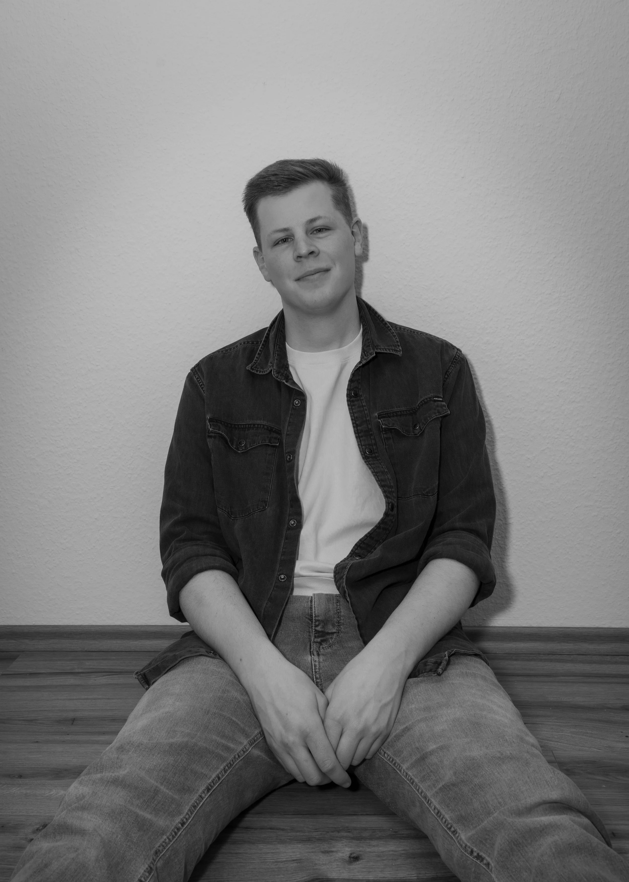 Ein junges, lächelndes männliches Model in einem schwarzen Jeansjacke und weißen T-Shirt sitzt auf dem Boden vor einer weißen Wand.