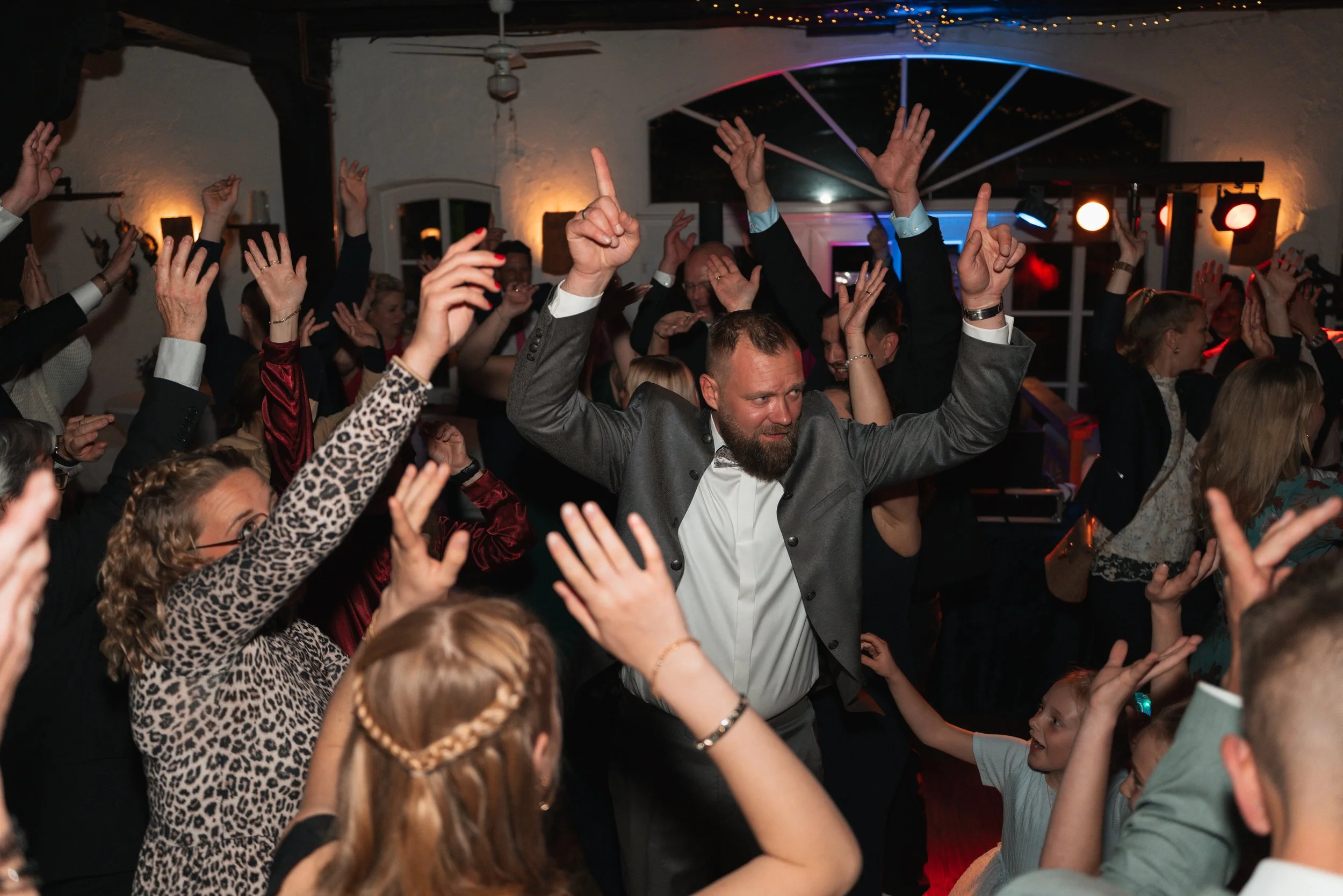 Menschen tanzen und feiern bei einer Party in einem festlich dekorierten Raum mit Lichterketten, bunten Lichtakzenten und einem DJ-Pult im Hintergrund.