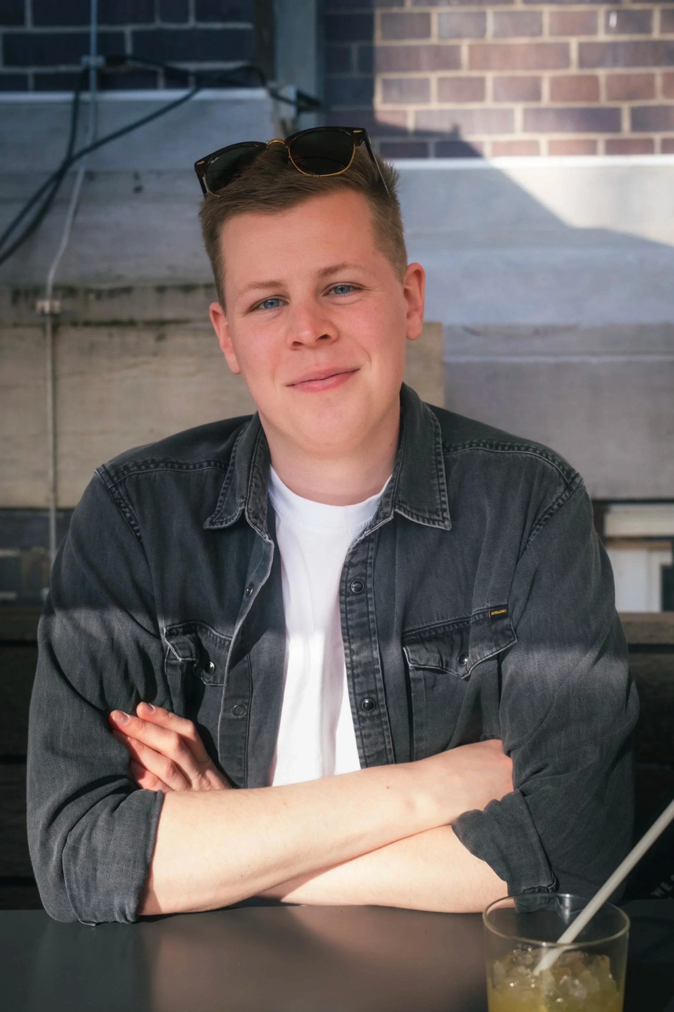 Junger Mann mit Sonnenbrille auf dem Kopf, lächelnd, sitzend an einem Tisch in einer gemütlichen, modernen Bar oder Café, mit einem Getränk mit Strohhalm vor sich.