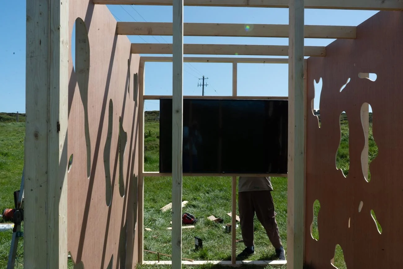 Under construction wooden structure with cutout designs, set outdoors on grassy field, with a person working inside and a television mounted on a wall.
