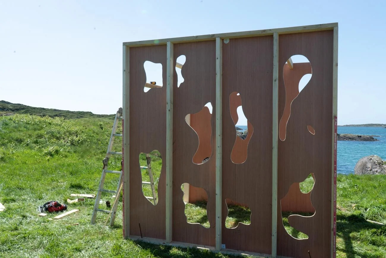 Wooden panel with irregular cut-out holes set up outdoors on grass near the coast, with a ladder and tools nearby