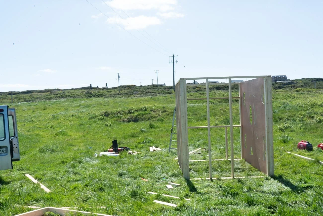 Construction site on a grassy field with a partially built wooden frame structure and tools scattered around, including a ladder, power tools, and wooden planks, under a bright sky.