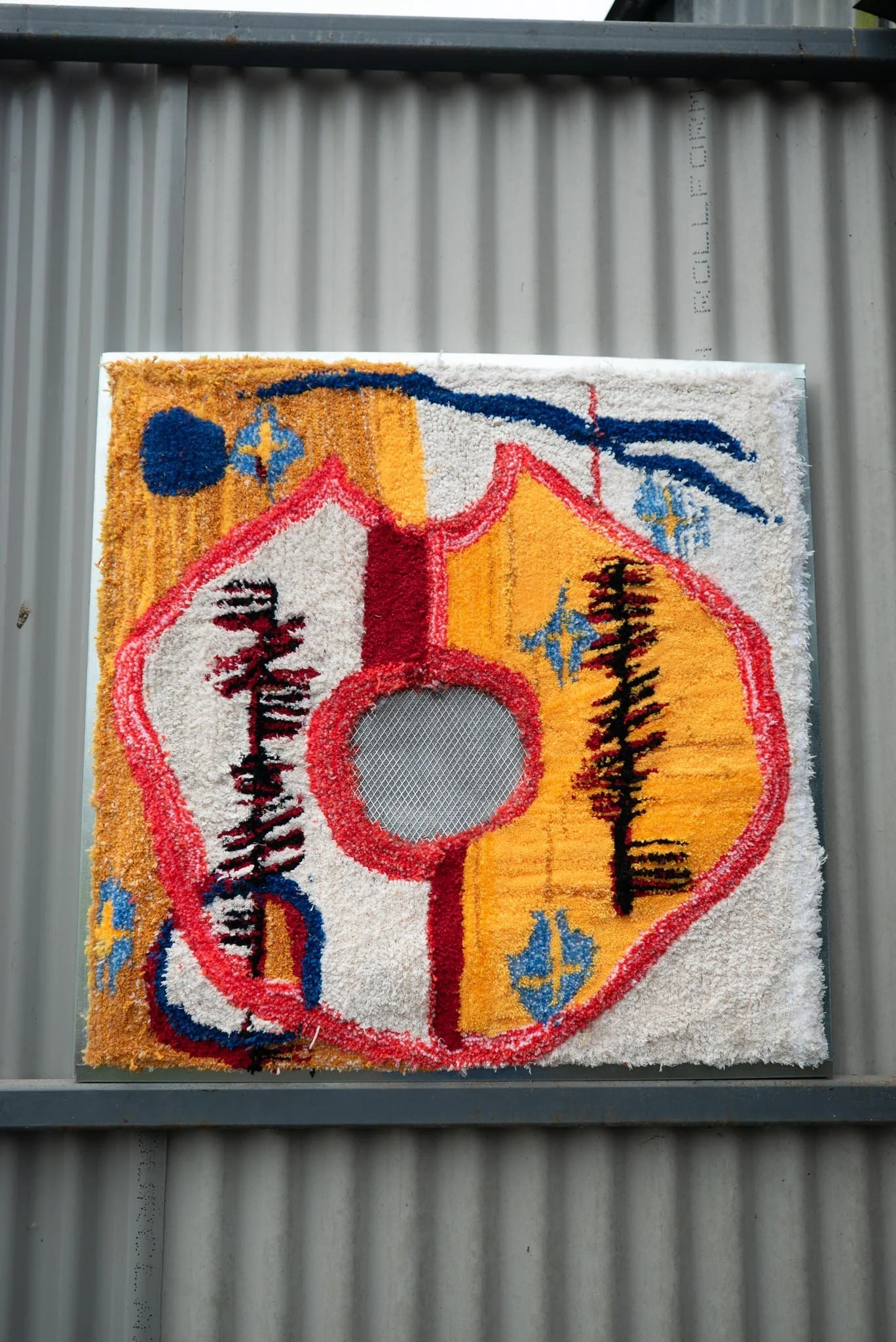 Close-up of a colorful, abstract woven wall hanging or rug with various shapes and patterns, mounted on a gray metal wall.