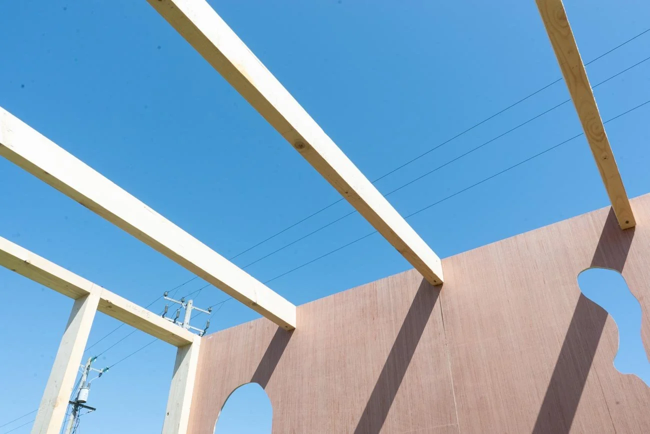 Partially constructed wooden structure with vertical and horizontal beams against a clear blue sky, with utility poles and power lines in the background.