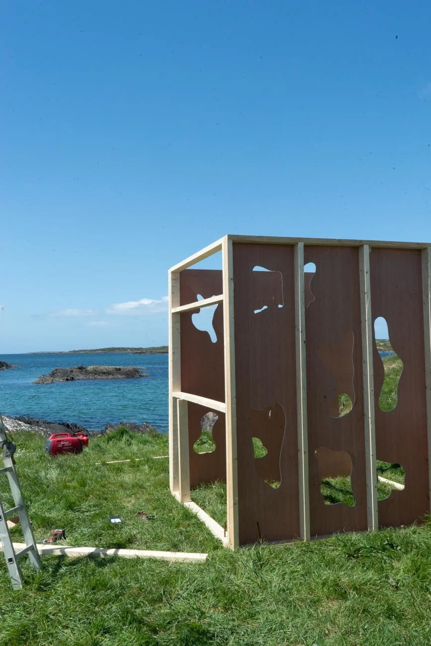 A partly constructed wooden art installation or sculpture on a grassy area by the coast, with the sea and a blue sky in the background.