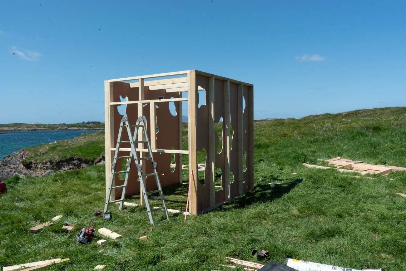 A partially constructed wooden structure on a grassy area next to a body of water, with tools and wood pieces scattered around. Art Installation, Sherkin Island for voice rewilding.