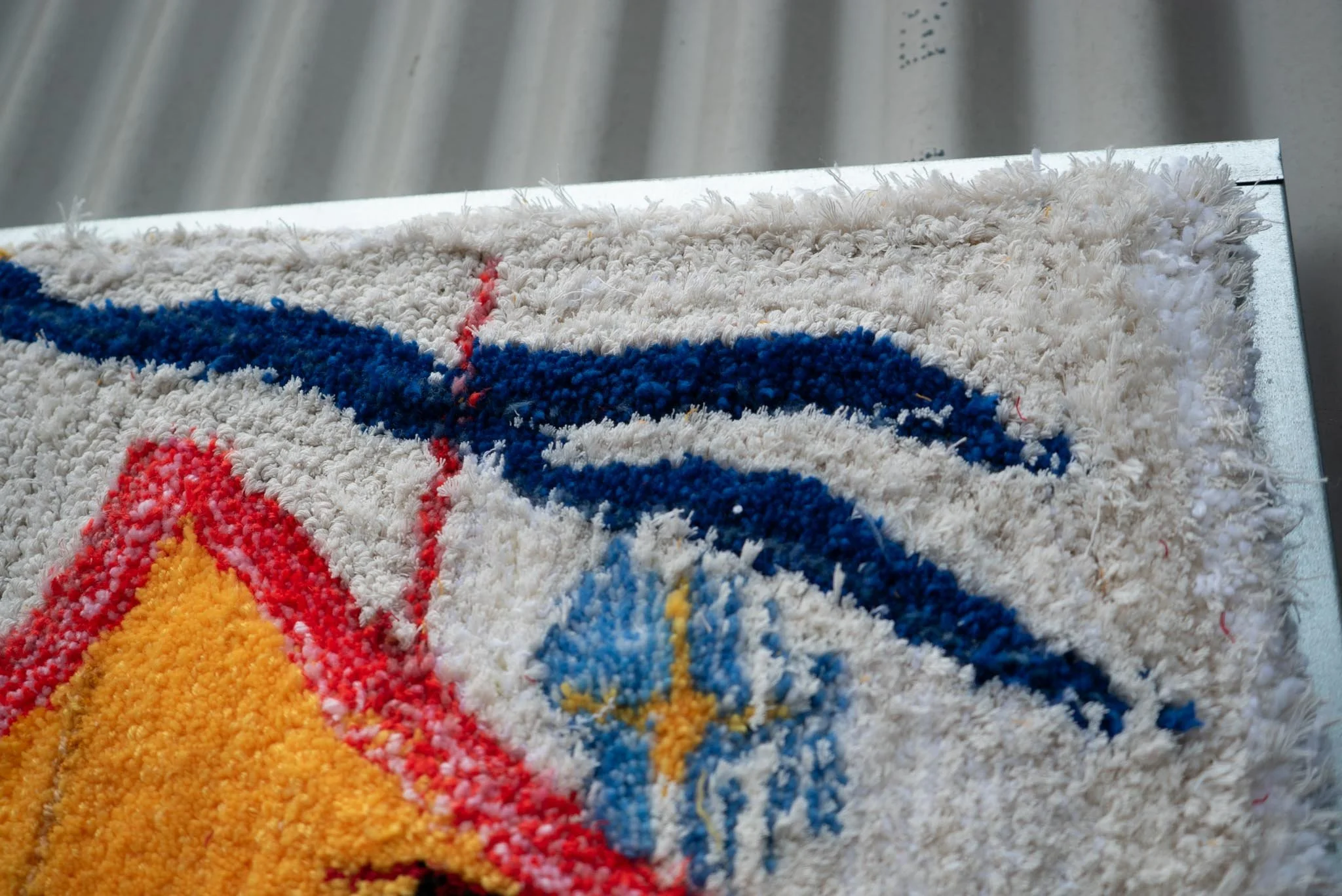 Close-up of a colorful textured carpet with a gold and red design on a shiny surface, background shows blurred vertical metallic panels.