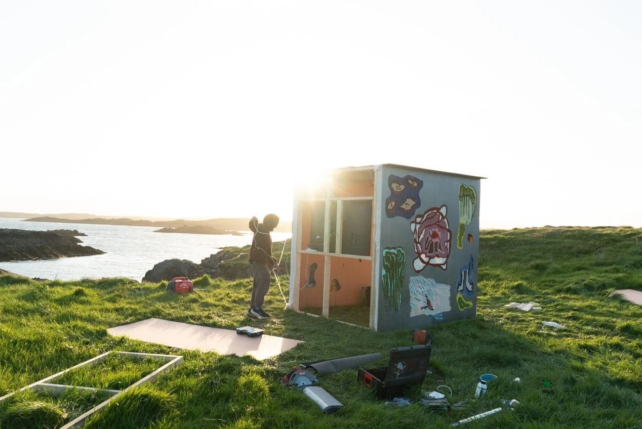 A person painting a small outdoor structure with colorful artwork at a coastal location during sunset, with grassy terrain and the ocean in the background.