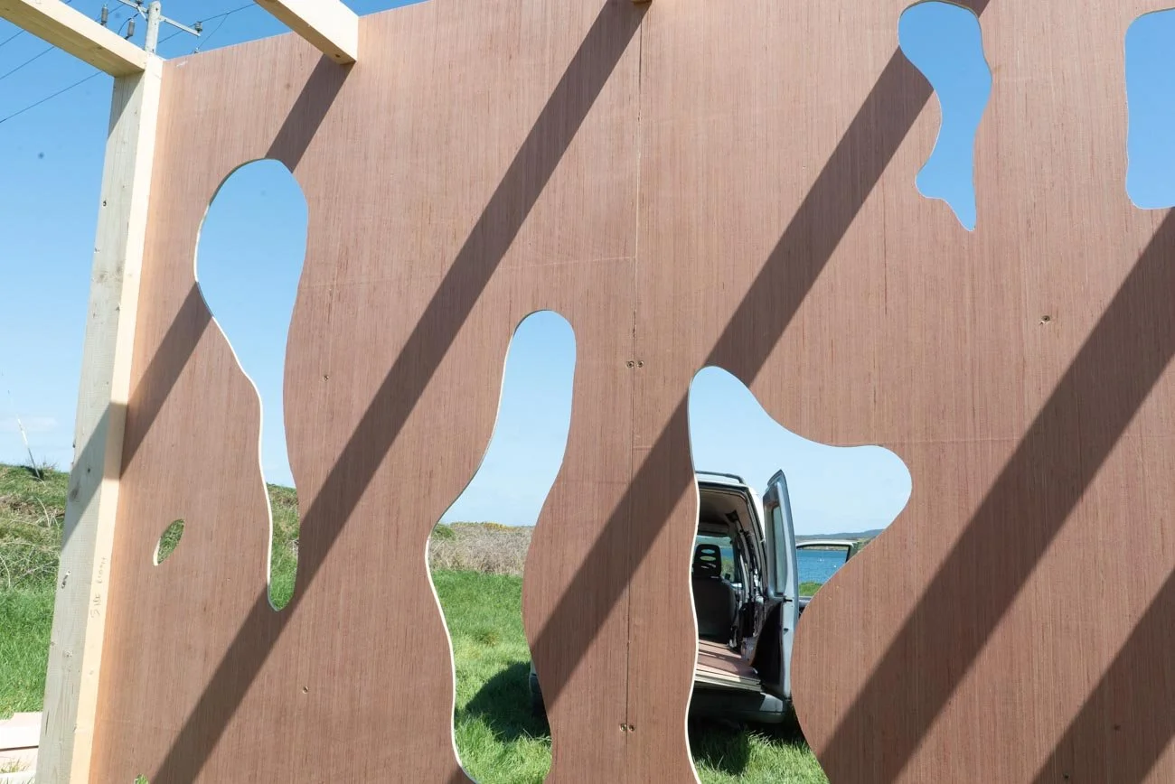 A wooden wall with irregularly shaped cutouts looks out onto a grassy area and a clear blue sky, with a vehicle visible through one cutout.