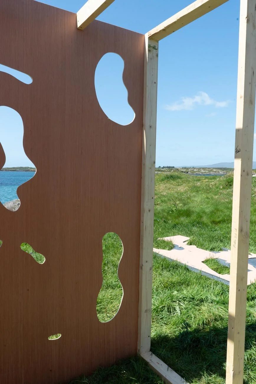 Partially constructed wooden frame and decorative panel with irregular cutouts, set outdoors near a grassy area and coastline under a blue sky.