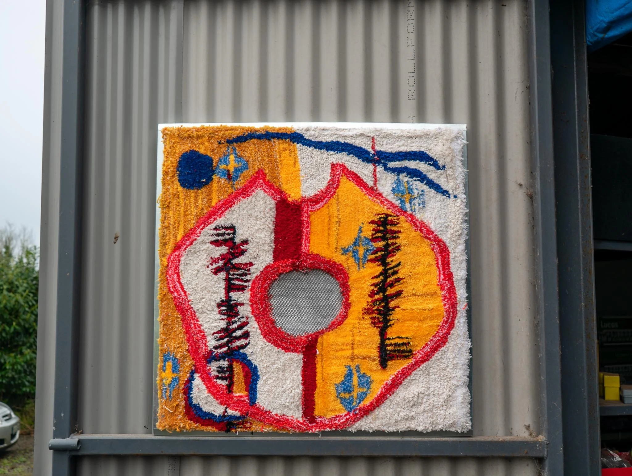 Colorful, patterned rug hanging on a metallic board outside, with trees and a cloudy sky in the background.