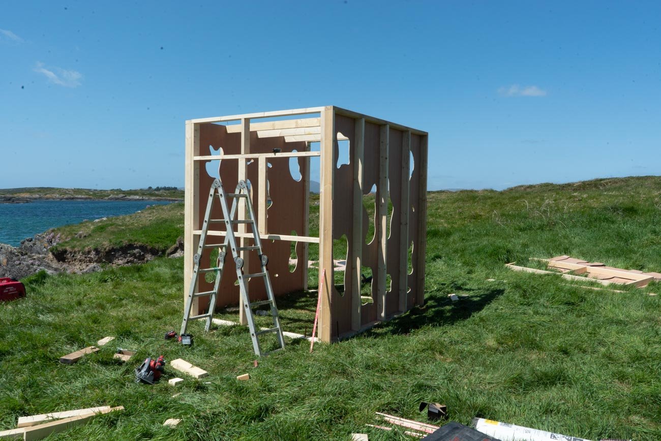 A partially constructed wooden shed on a grassy hill near the coast, with tools and building materials scattered around.