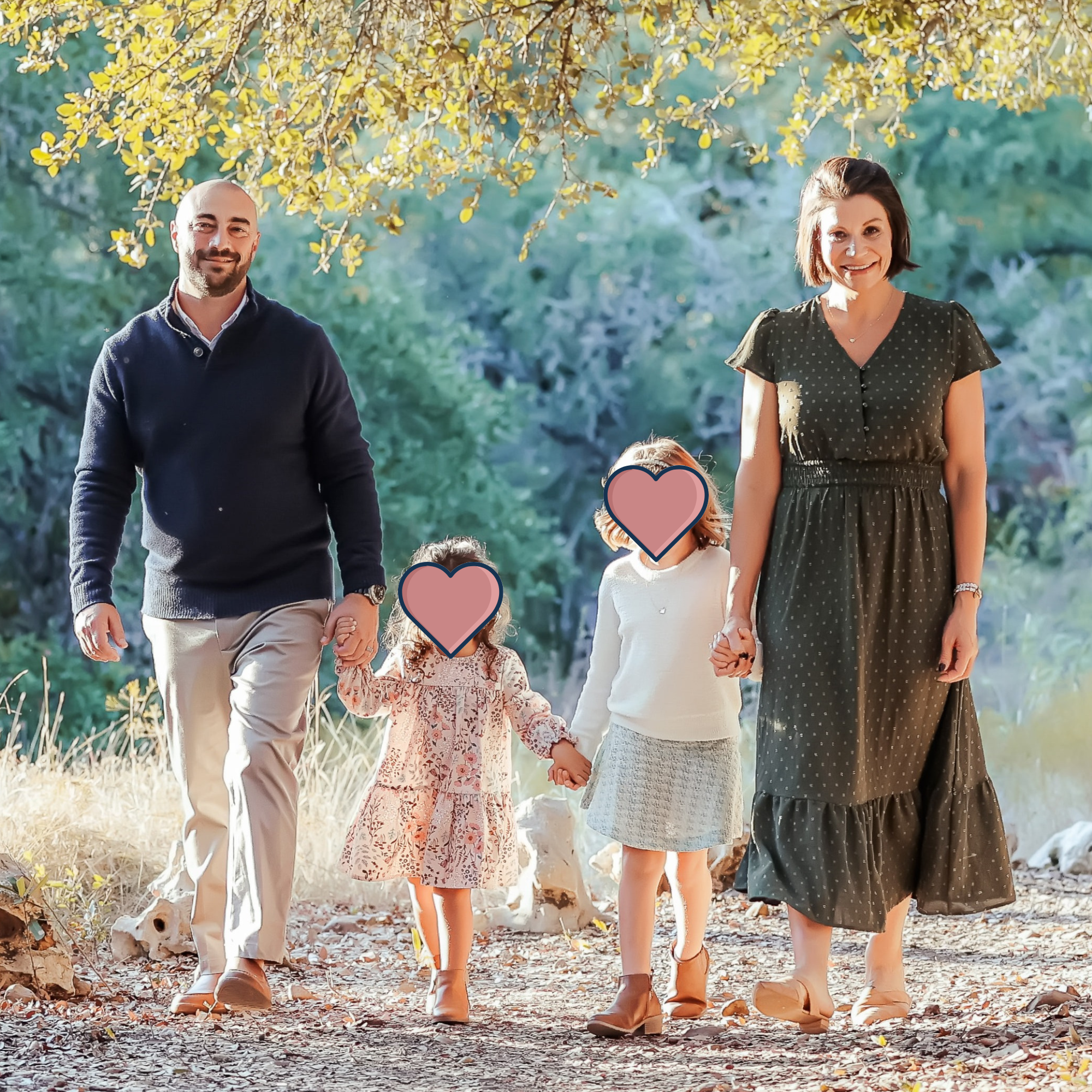 A family walking outdoors in a park during fall. The family includes a man, a woman, and three children, with two children holding hands. The man is wearing a navy sweater and beige pants, the woman is in a dark green polka dot dress, and the children are in light-colored outfits. The background features trees with yellow leaves and natural scenery.