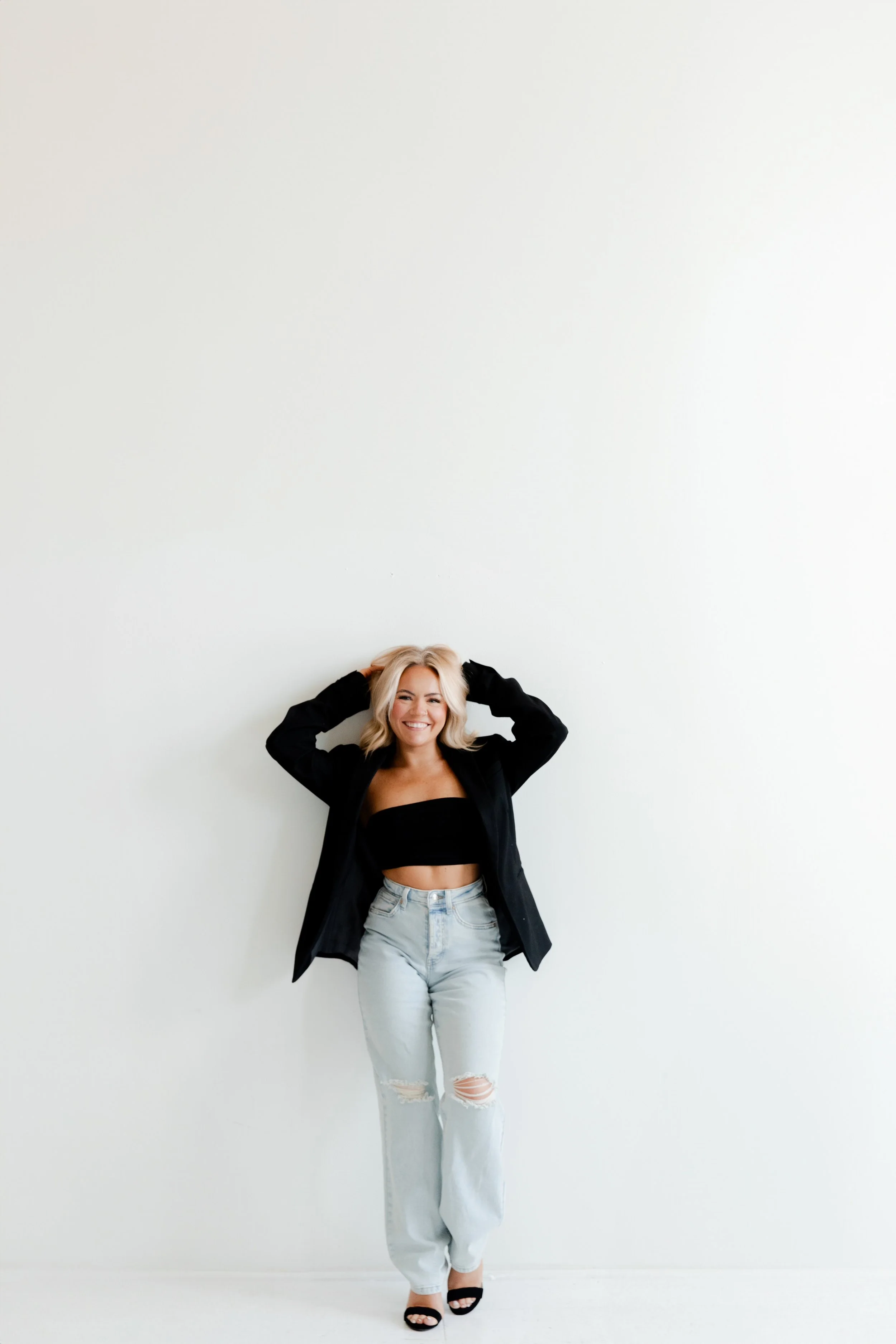 A woman with blonde hair wearing a black crop top, light blue ripped jeans, black heels, and a black blazer, standing against a plain white wall, smiling with her hands in her hair.