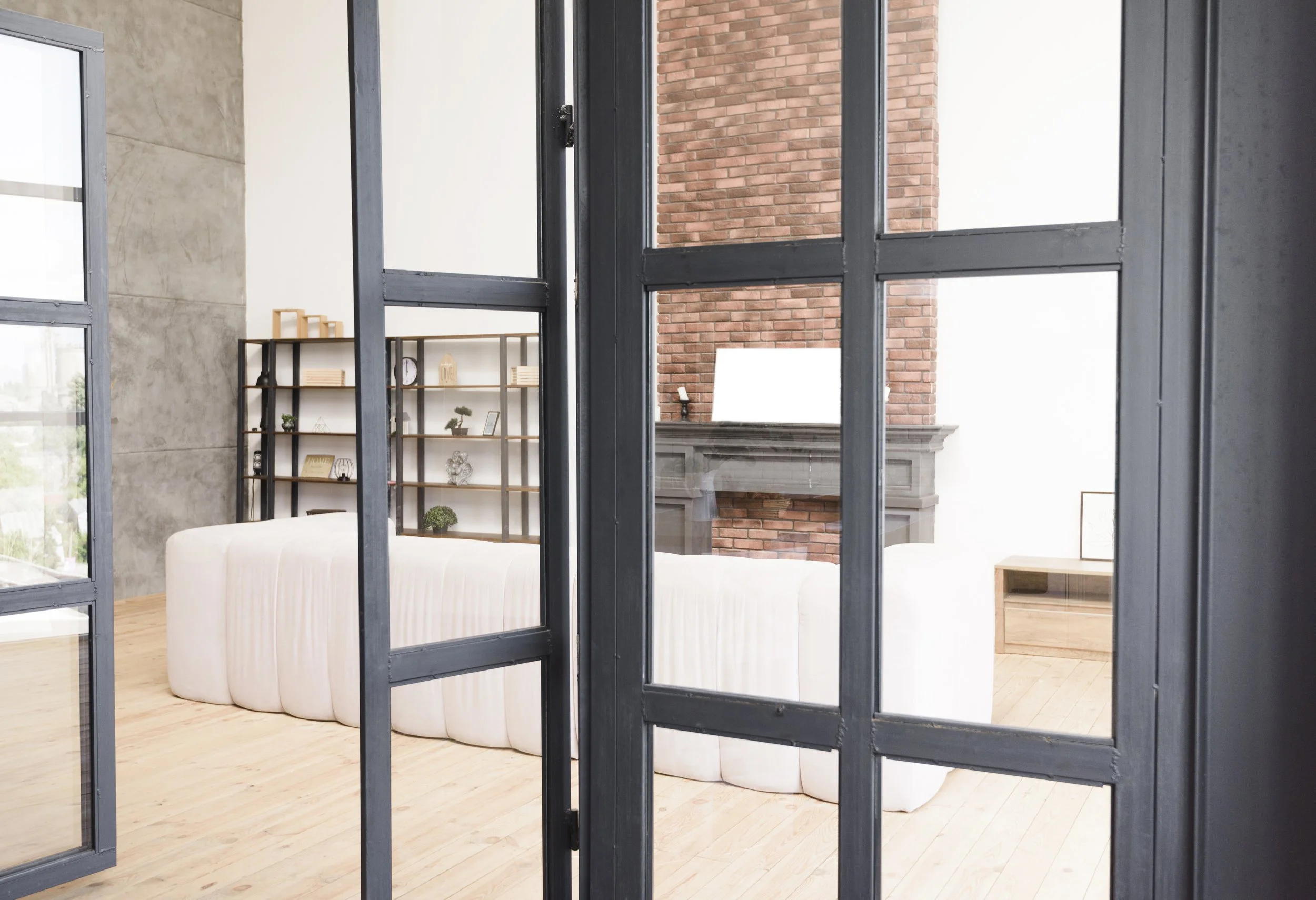 Interior view of a modern living room seen through black-framed glass doors, featuring a white sofa, a wooden bookshelf, a brick fireplace, and a wooden floor.