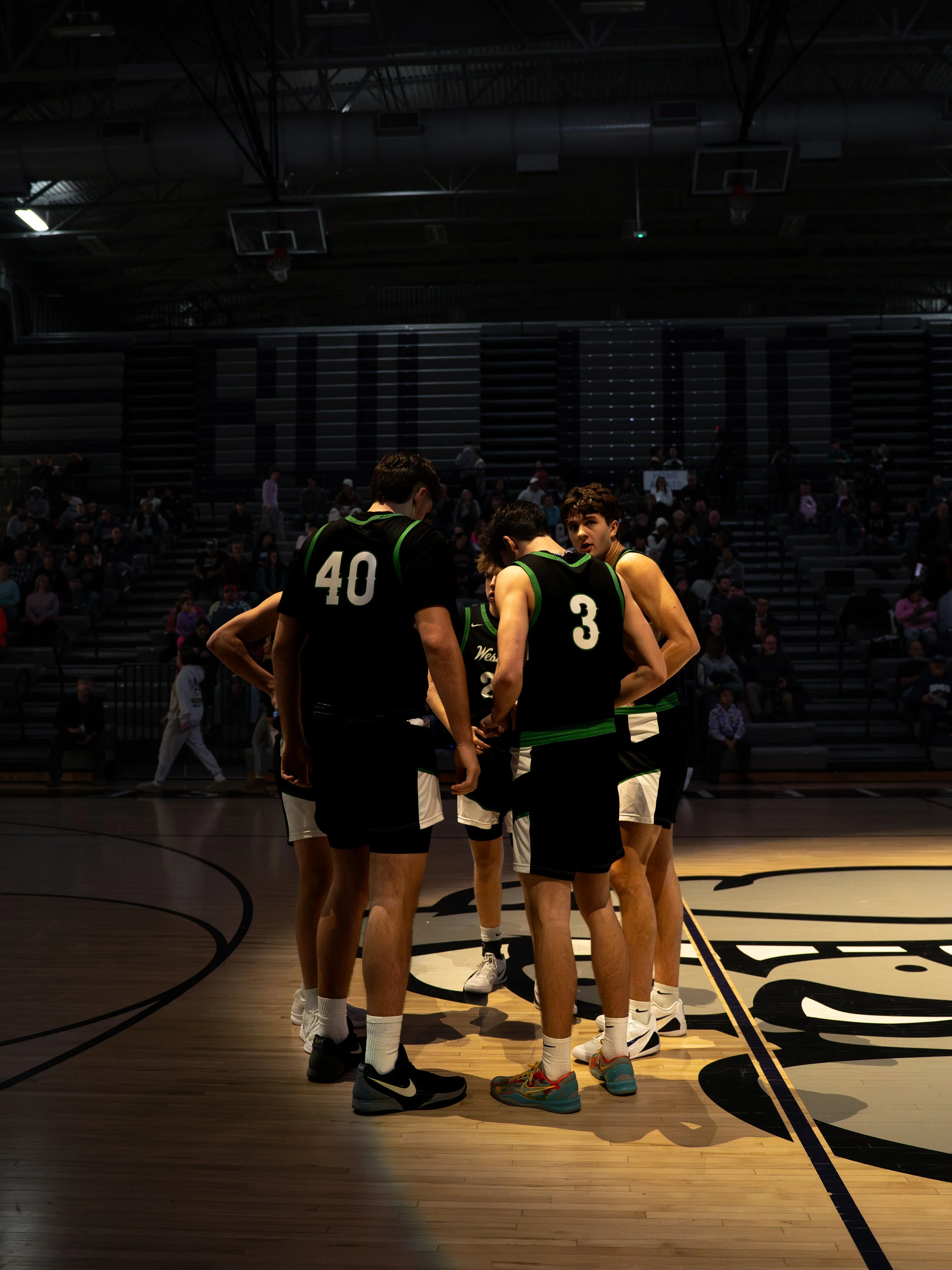 A group of basketball players in black and green uniforms huddle on a basketball court during a game or practice, with spectators seated in the background.