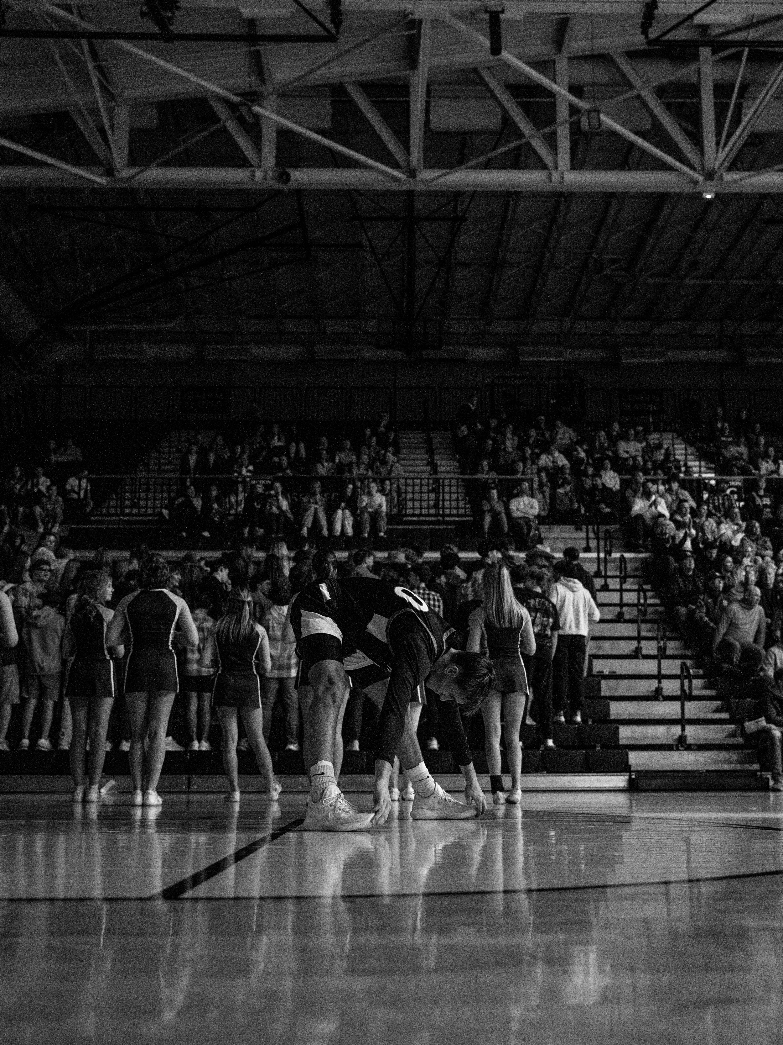 A volleyball player is tying their shoe on a gymnasium court with an audience watching from the bleachers.
