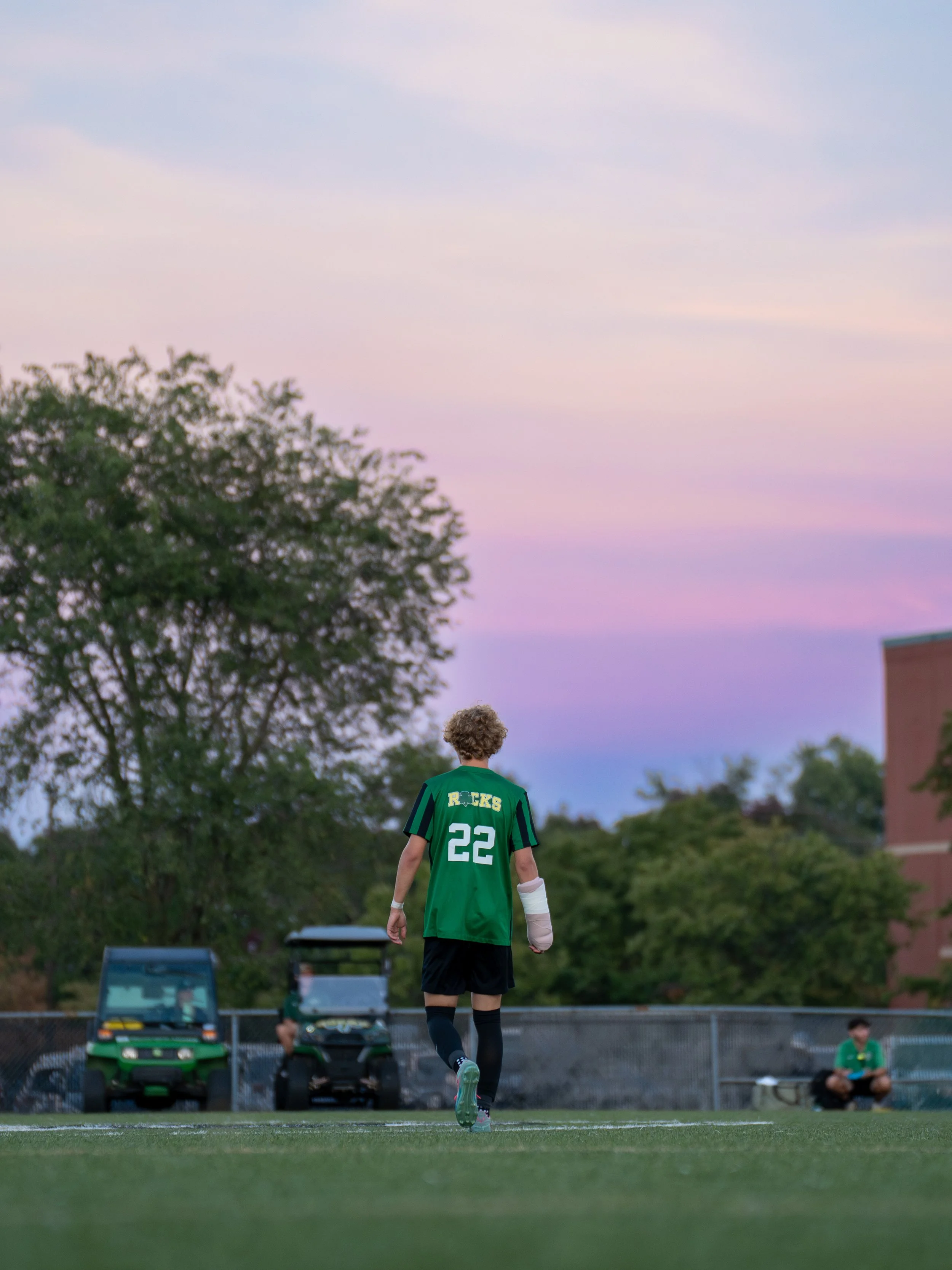 A soccer player with curly hair, wearing a green jersey with the number 22 on the back and black shorts, walking on a soccer field during sunset, with a few people sitting on the sidelines and trees in the background.