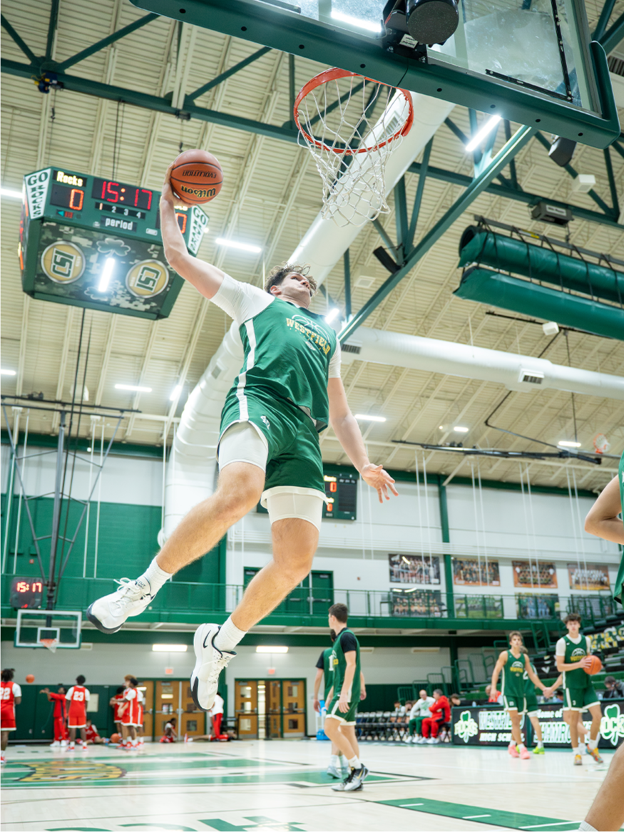 A basketball player in a green jersey and white shorts jumps to dunk a basketball in a gymnasium during a game, with players on the court and scoreboard visible.