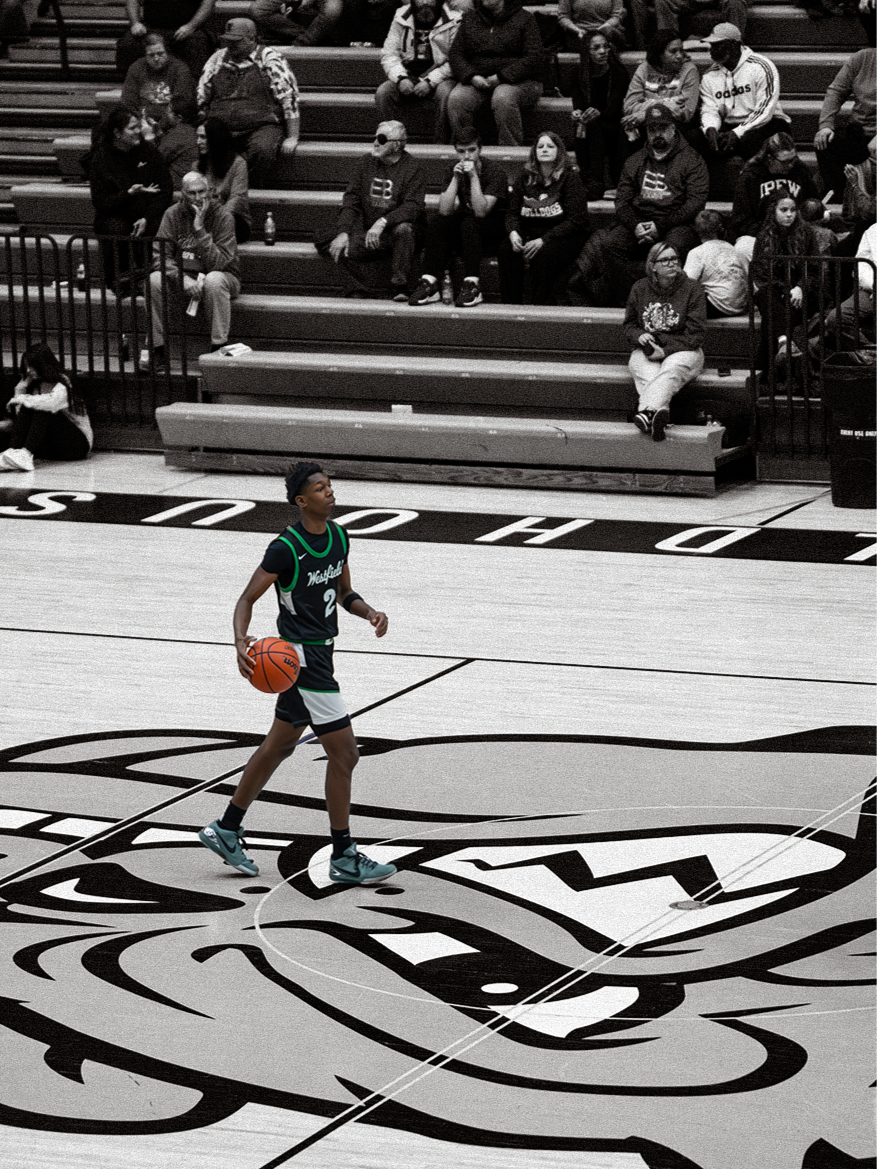 A young basketball player dribbling a basketball on an indoor court with an audience seated on bleachers in the background.