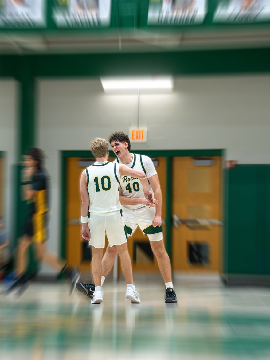Two basketball players celebrating on the court, one wearing jersey number 10 and the other wearing jersey number 40, both in white uniforms with green accents, in a gymnasium with green walls and an exit door behind them.