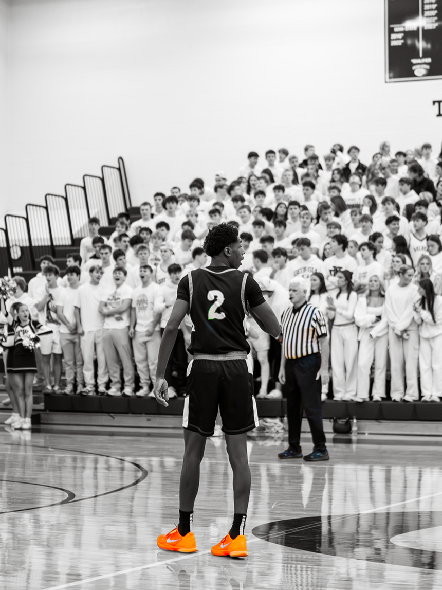 A basketball player wearing a black jersey with the number 2 and bright orange shoes standing on the court, with a crowd of students in the bleachers in the background.