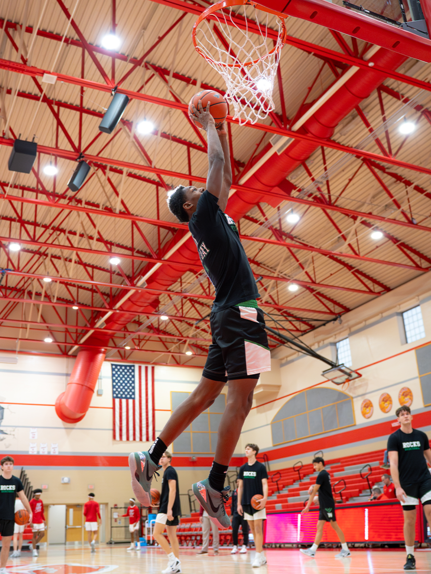 A basketball player is jumping to make a shot in a gymnasium, with other players and coaches watching and practicing in the background.